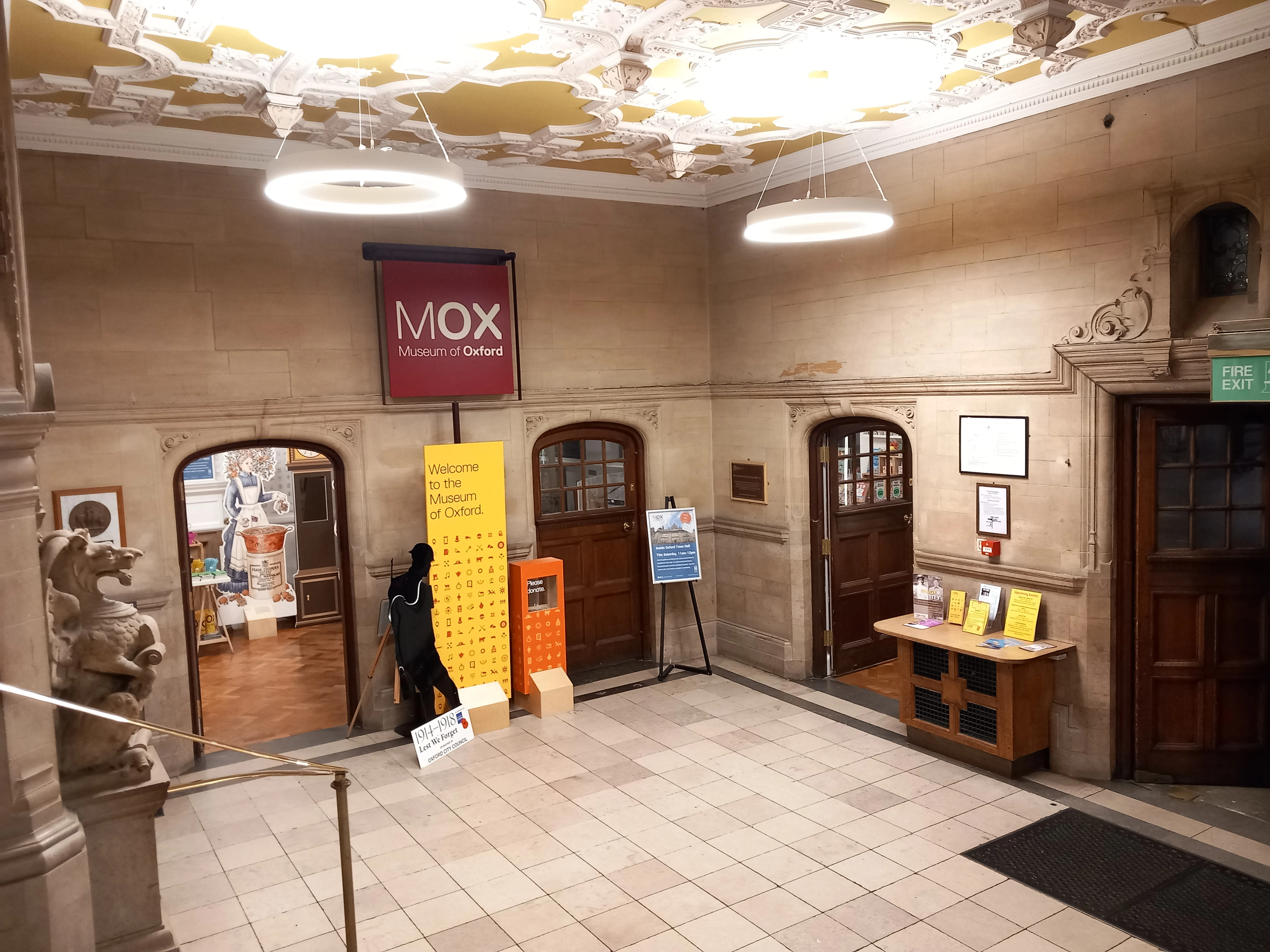 Vestíbulo interior del Museum of Oxford con un letrero colgante de MOX, puertas arqueadas, paredes de piedra, suelo de baldosas y un techo decorativo con lámparas circulares