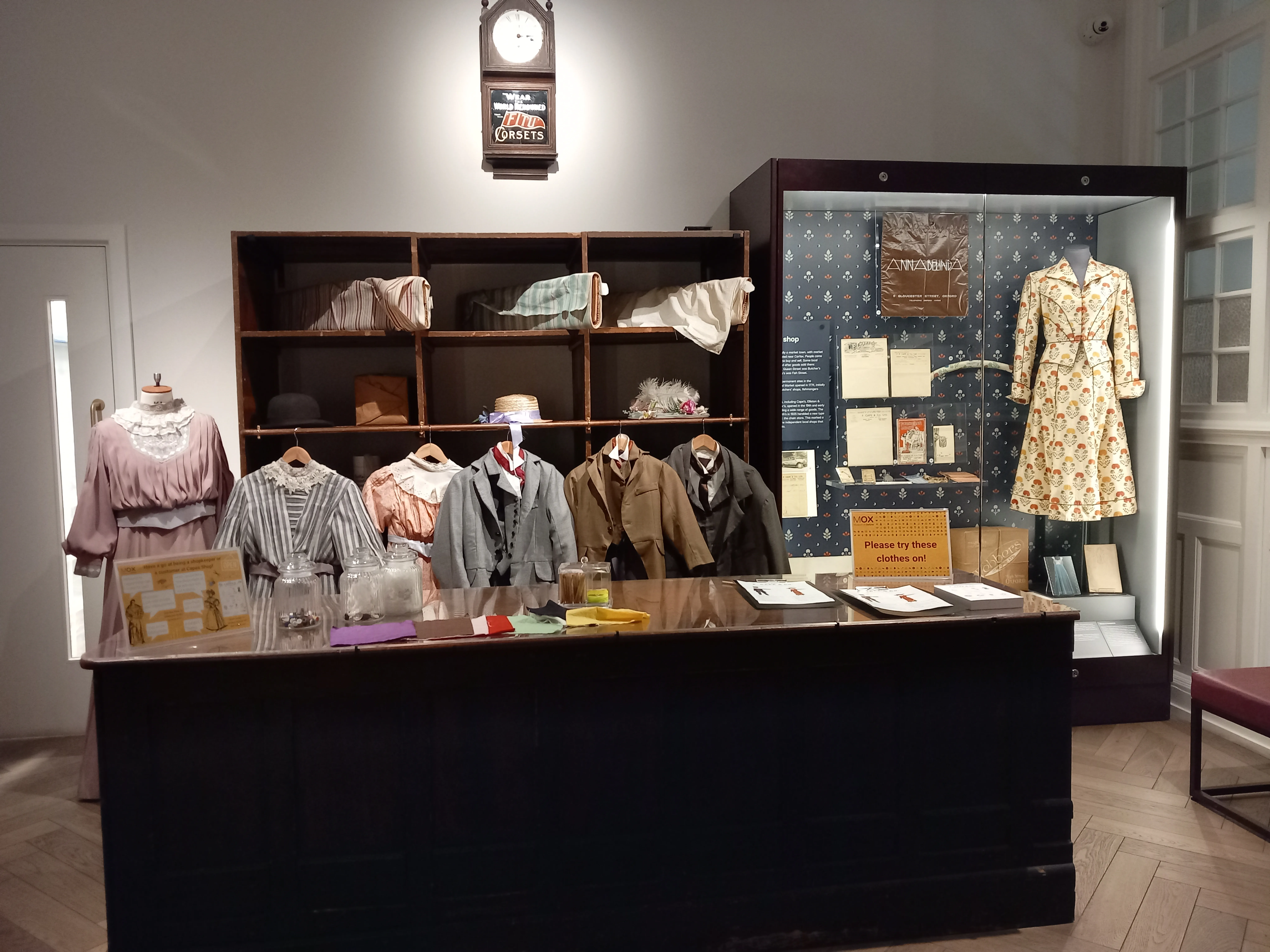 Exhibición en el Museum of Oxford que muestra ropa histórica en maniquíes y perchas, estanterías con artefactos y una vitrina de cristal con documentos y un vestido de flores