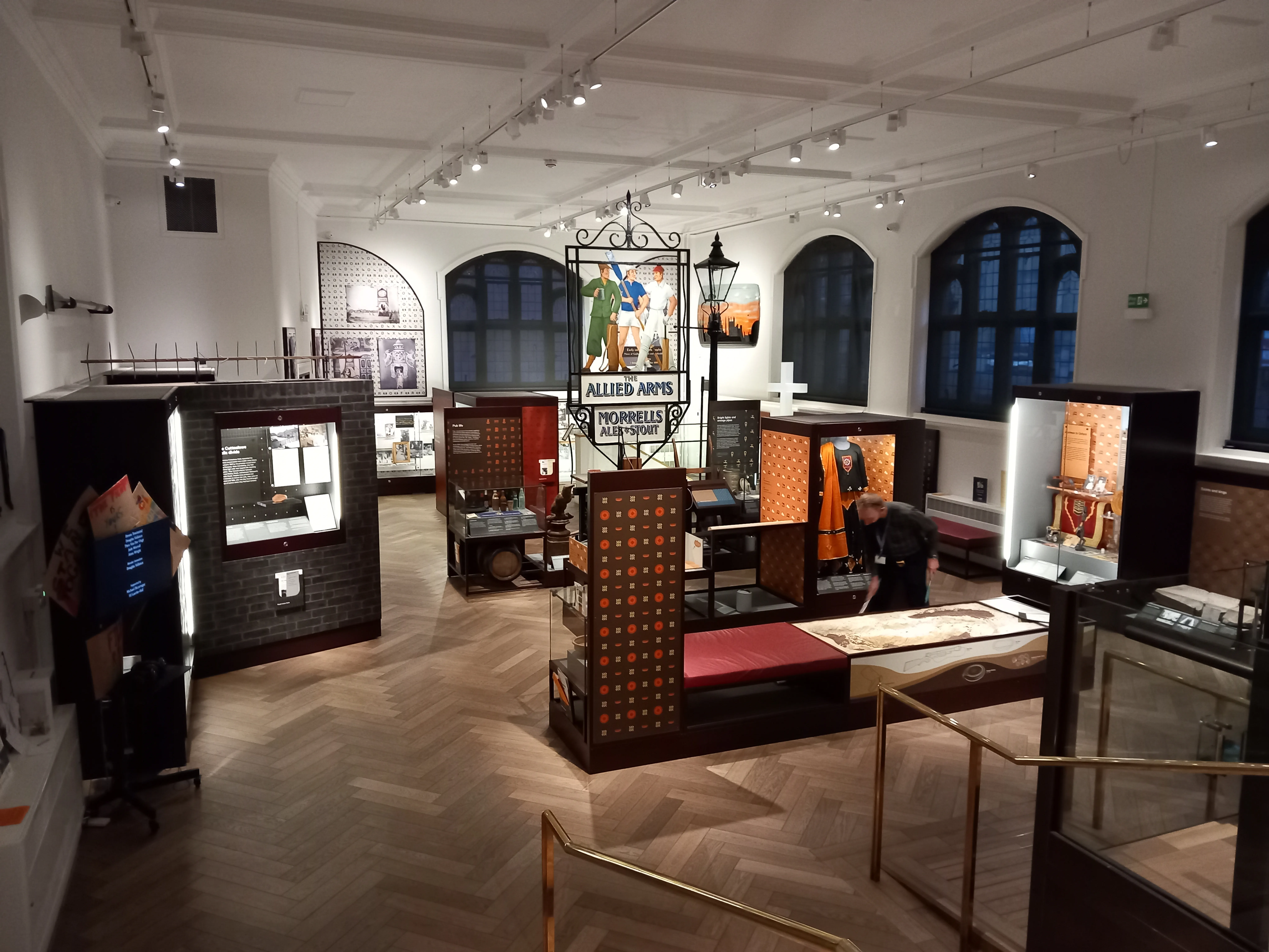 Vista interior amplia de una galería de museo con suelos de madera en espiga, vitrinas de madera oscura y un gran cartel colgante que dice 'Allied Armies' frente a ventanas arqueadas.