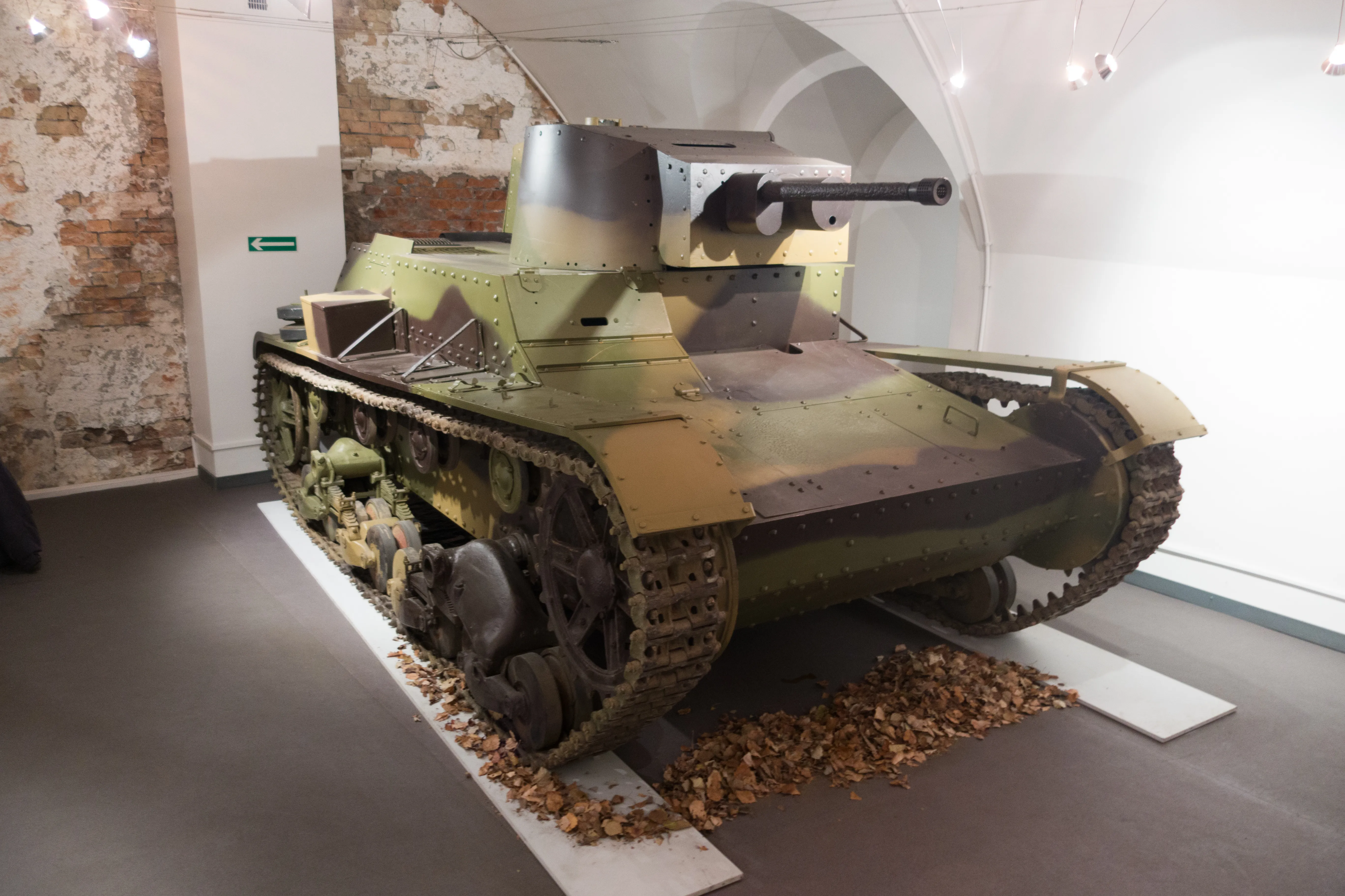 Een groene en bruine militaire tank tentoonstelling in een museumgalerij met booggewelven en zichtbare bakstenen muren.