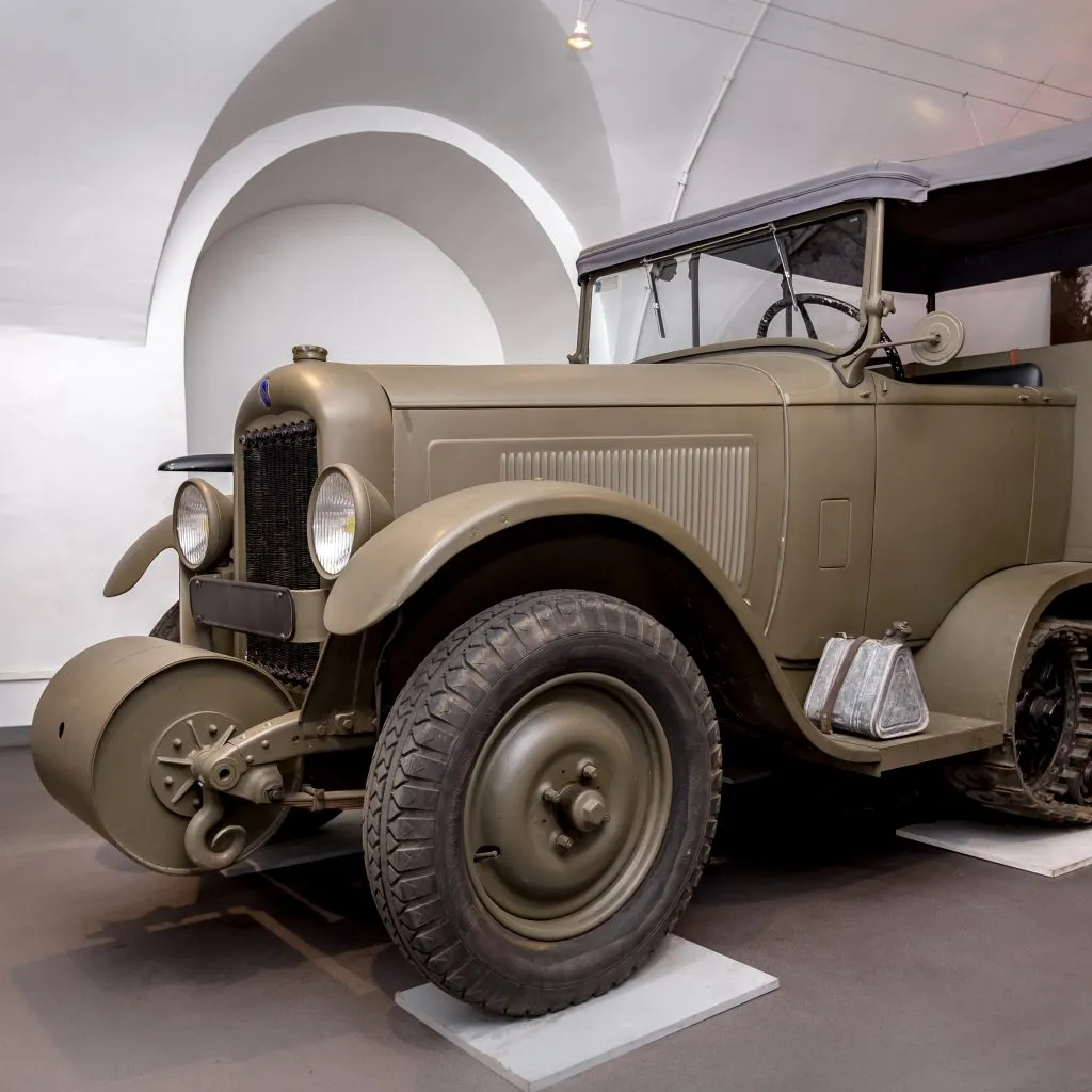 Een vintage olijfgroene militaire truck met grote wielen en een ronde spatbord, tentoongesteld binnenshuis onder witte boogplafonds.