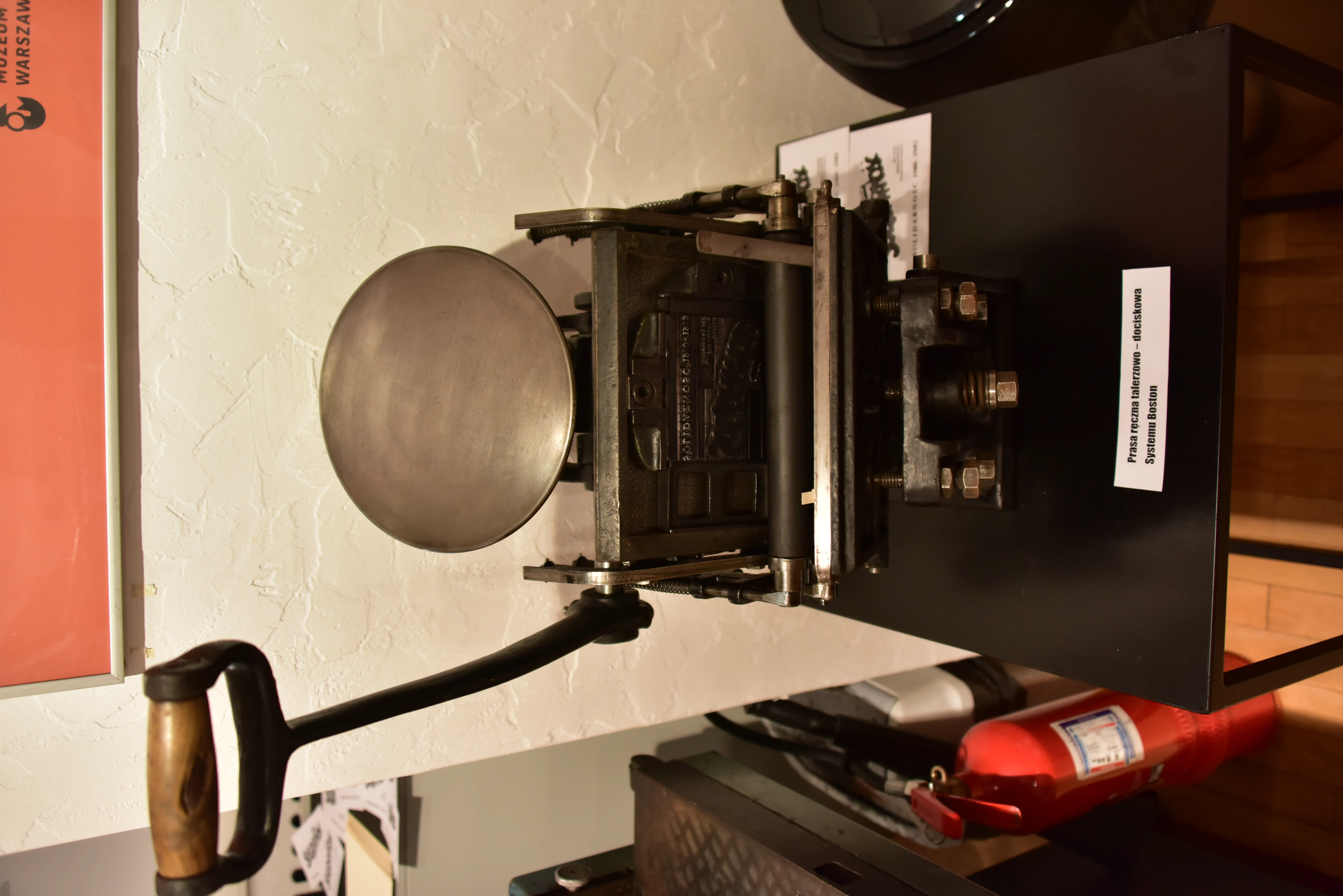 Een vintage drukpers met een grote ronde plaat en een hendel, tentoongesteld op een zwarte tafel met een informatief bord.