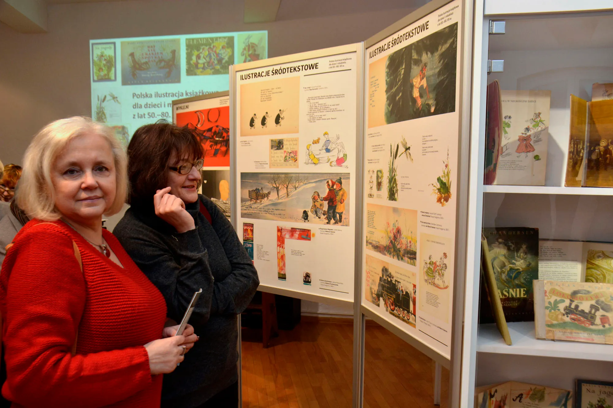 Twee vrouwen staan voor een tentoonstellingspaneel met illustraties en tekst over Poolse kinderboekillustraties.