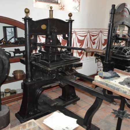 Een grote zwarte vintage drukpers met messing accenten en houten handvatten, tentoongesteld in een museumzaal met betegelde vloeren en historische artefacten.