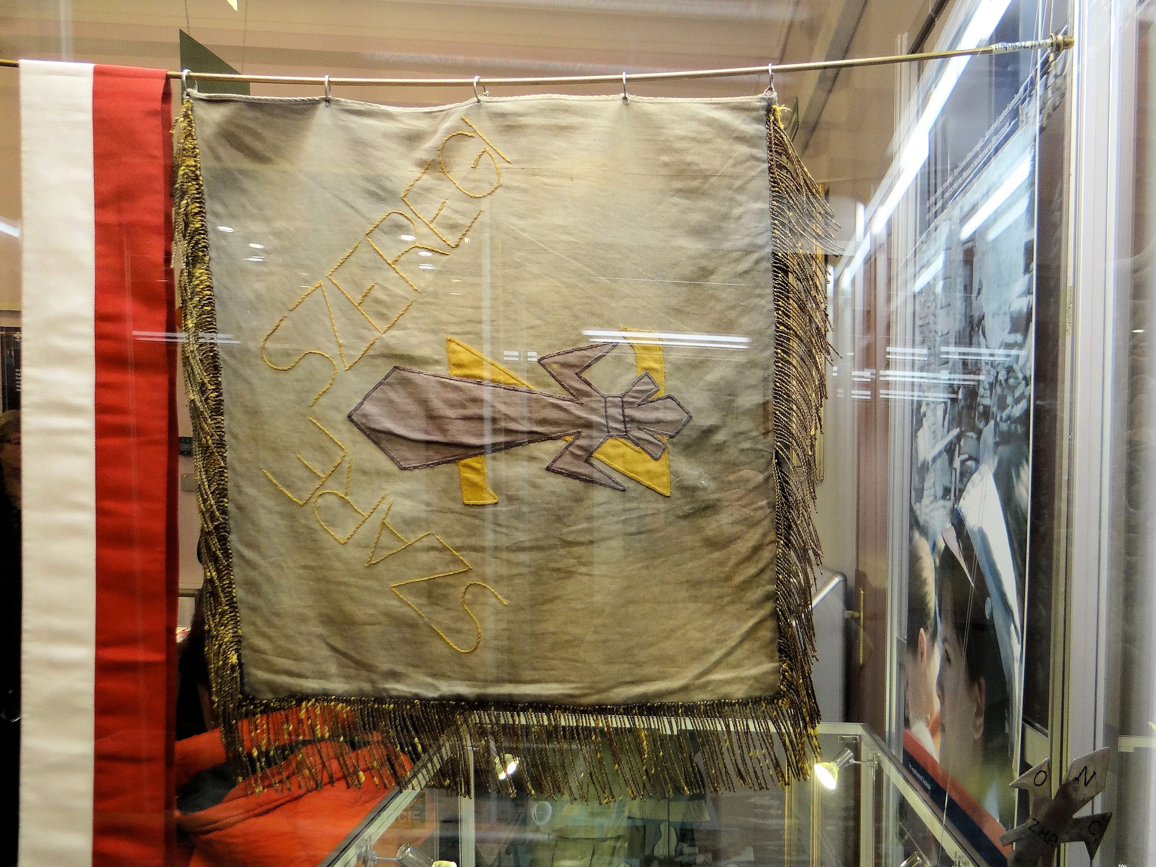 Geborduurd scoutingvaandel met een kruisontwerp en gefranjerde randen, tentoongesteld in een vitrine