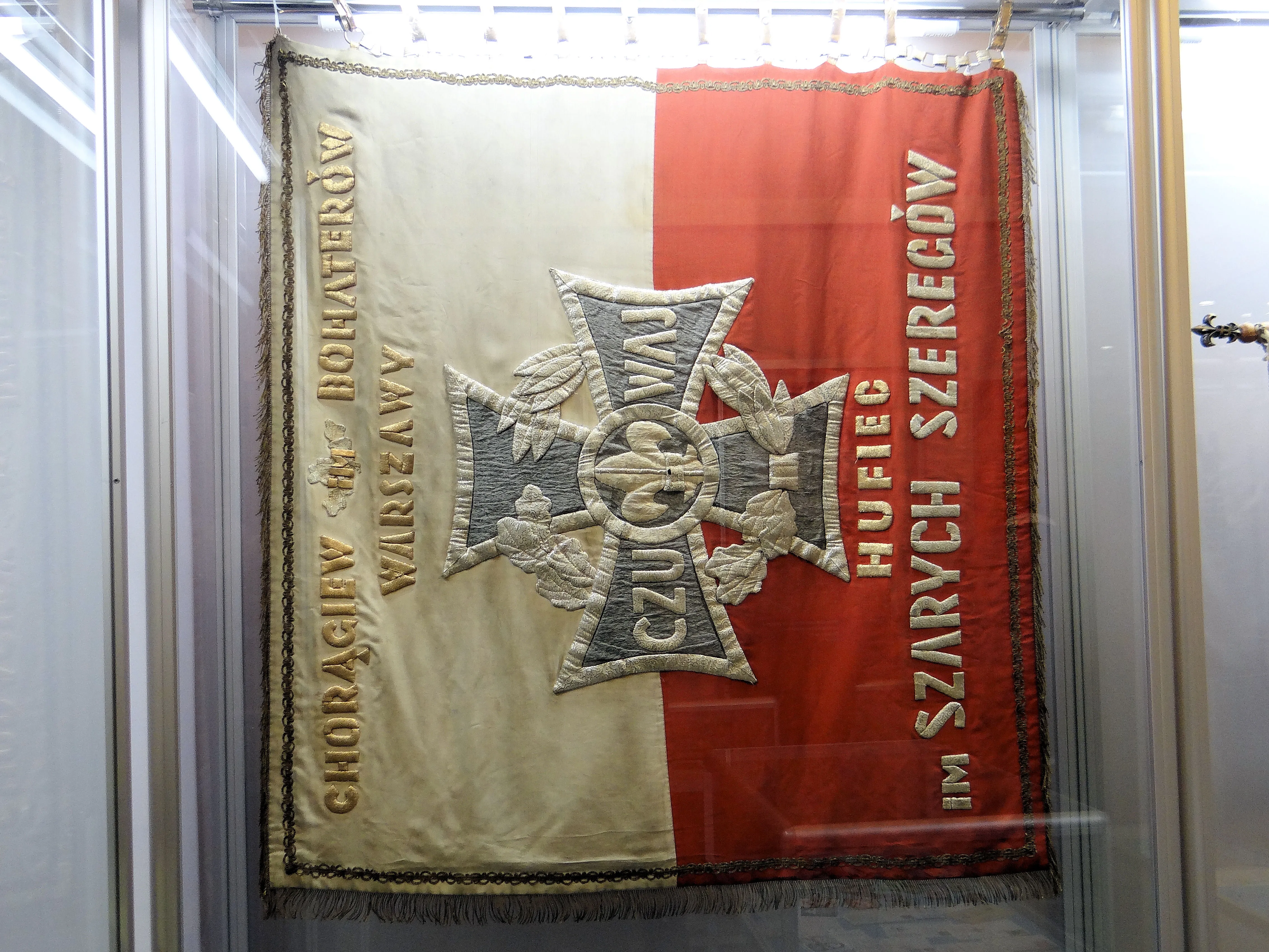 Een rood-witte scoutingvlag met een centraal kruisembleem en Poolse tekst, tentoongesteld in een vitrine.