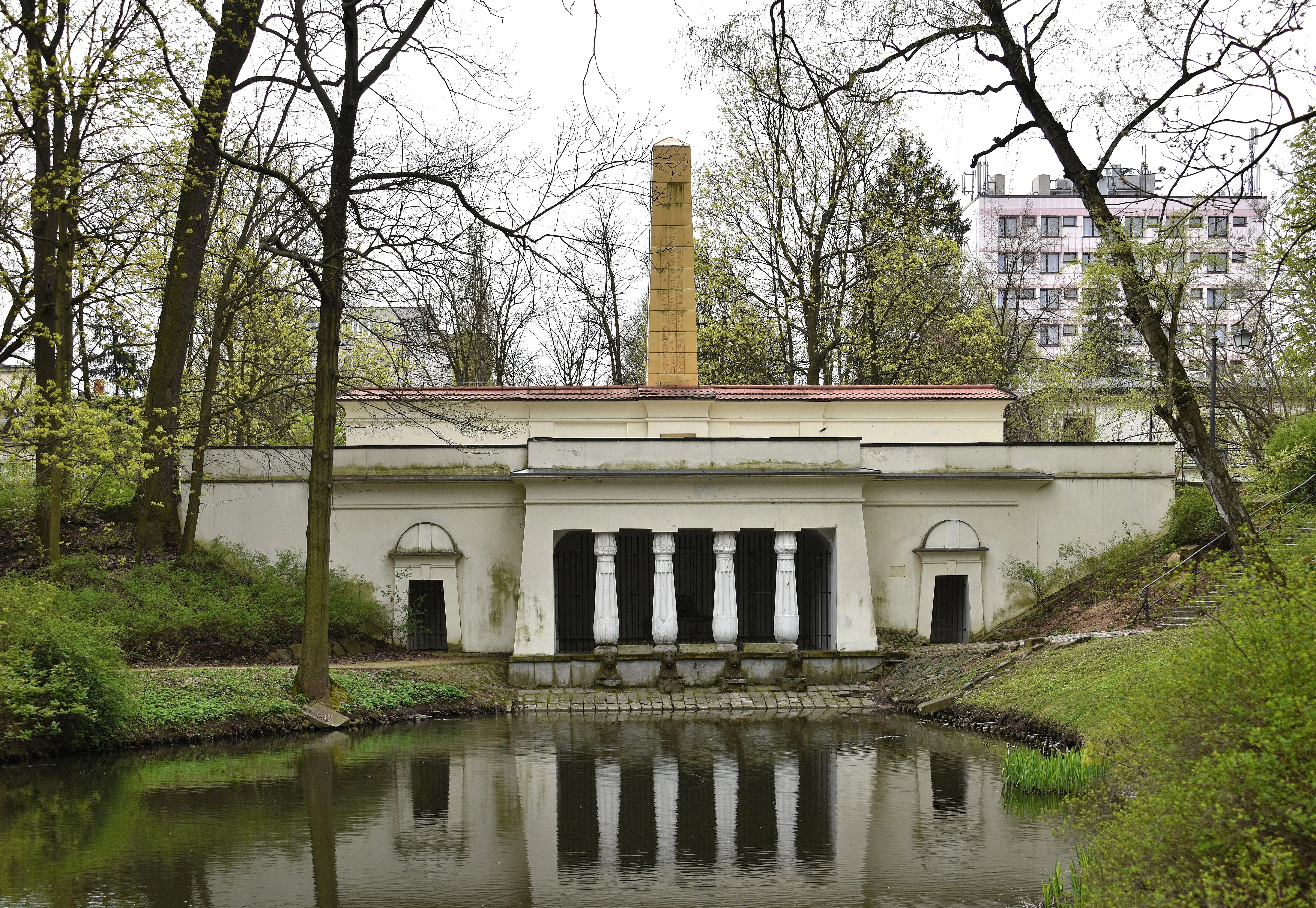 Neoklassiek gebouw met zuilen en een hoge gele schoorsteen die weerspiegeld wordt in een vijver, omringd door bomen en groen.