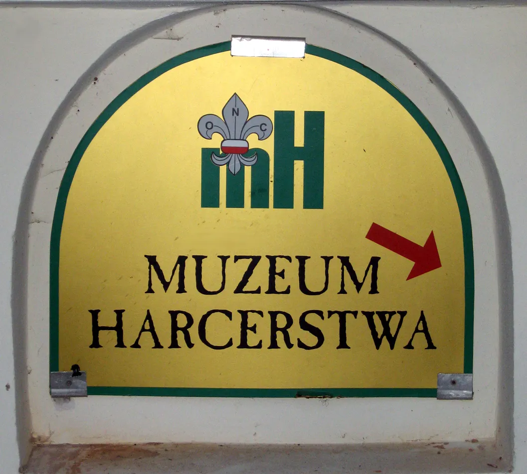 Gele boogvormige uithangbord met groene rand waarop het logo en de naam 'MUZEUM HARCERSTWA' van het Museum van Scouting staan, met een rode pijl naar rechts.