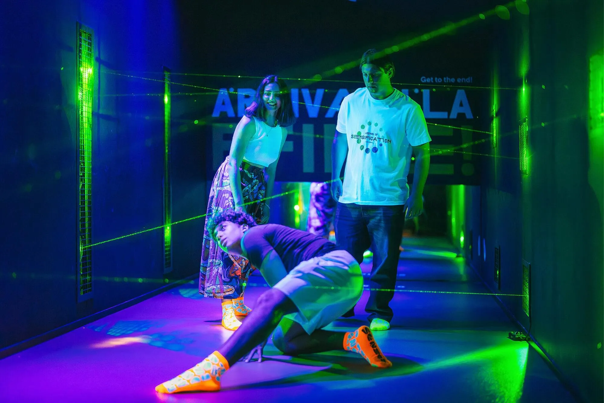 Trzy osoby w ciemnym pokoju z zielonymi i fioletowymi światłami laserowymi, wchodzące w interakcję z wystawą w Muzeum Zmysłów w Madrycie.