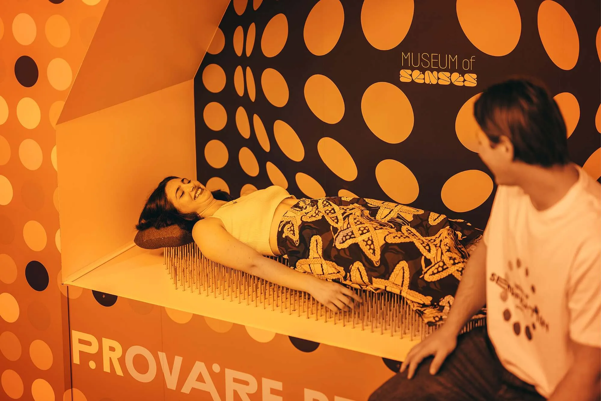 A person lying on a bed of nails in a room with polka-dot walls and the Museum of Senses logo