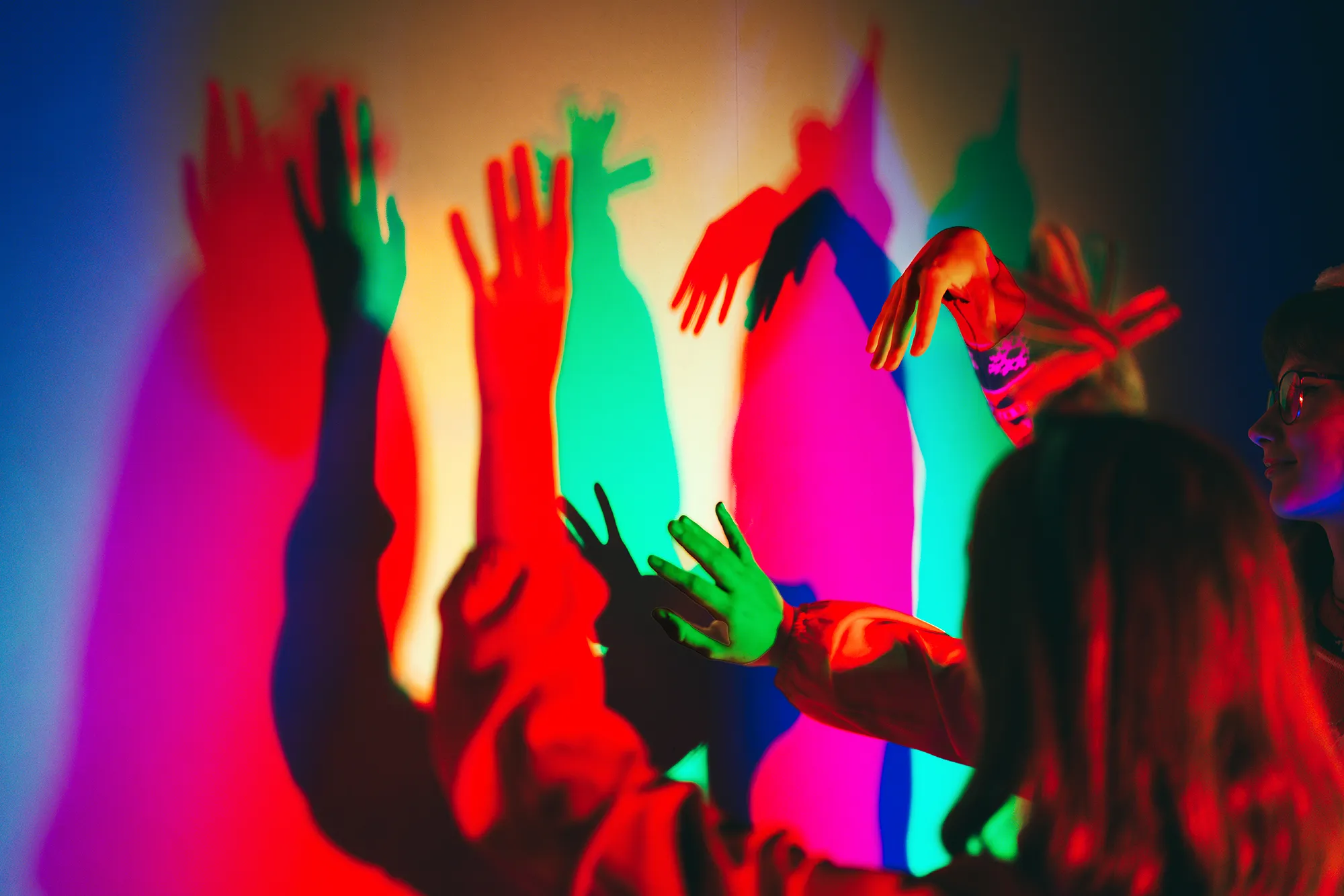 Hands reaching towards a wall with colorful light projections casting vibrant red, green, and blue shadows