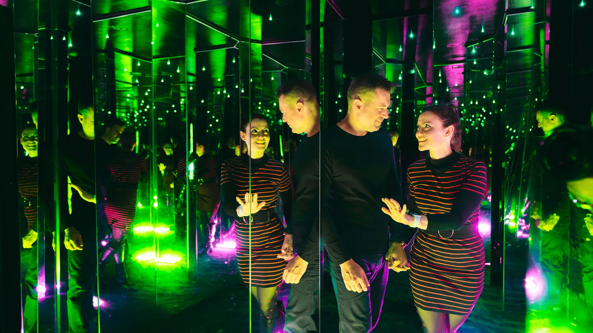 Two people standing in a mirrored room with green and purple lights creating a kaleidoscopic reflection effect