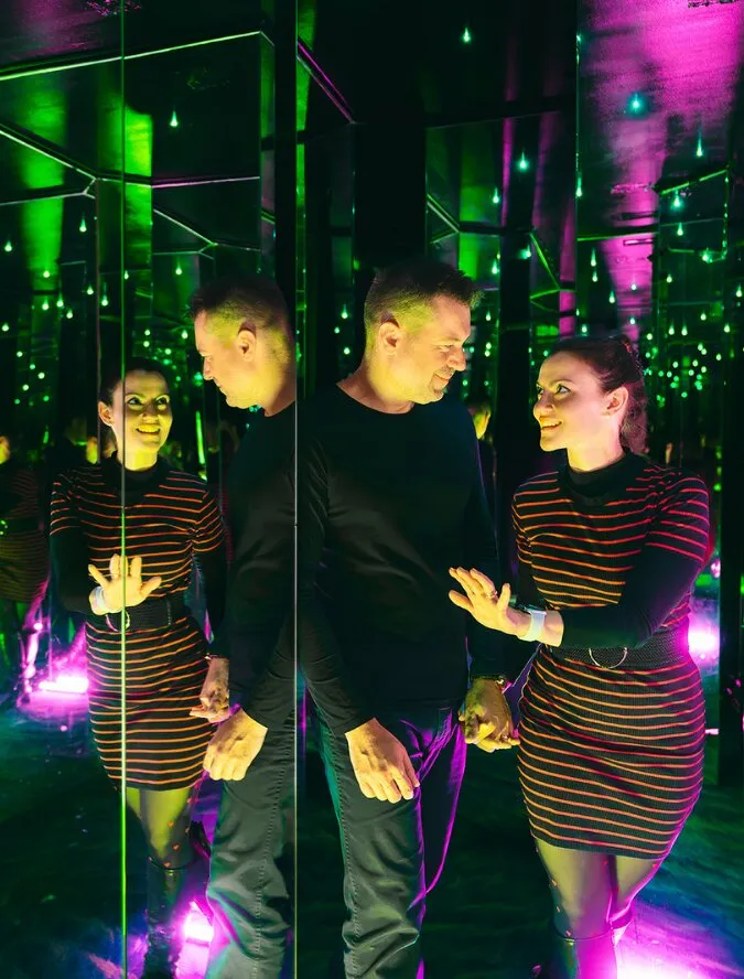 Two people standing in a mirrored room with green and purple lighting, surrounded by reflections and glowing dots.