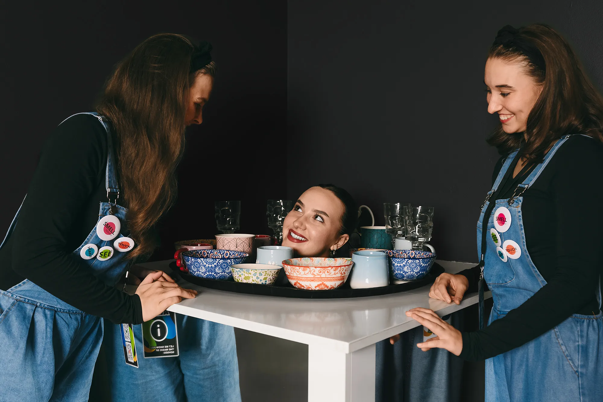 Two women in denim overalls standing around a table with bowls and glasses, one woman's head appearing to float above the table.