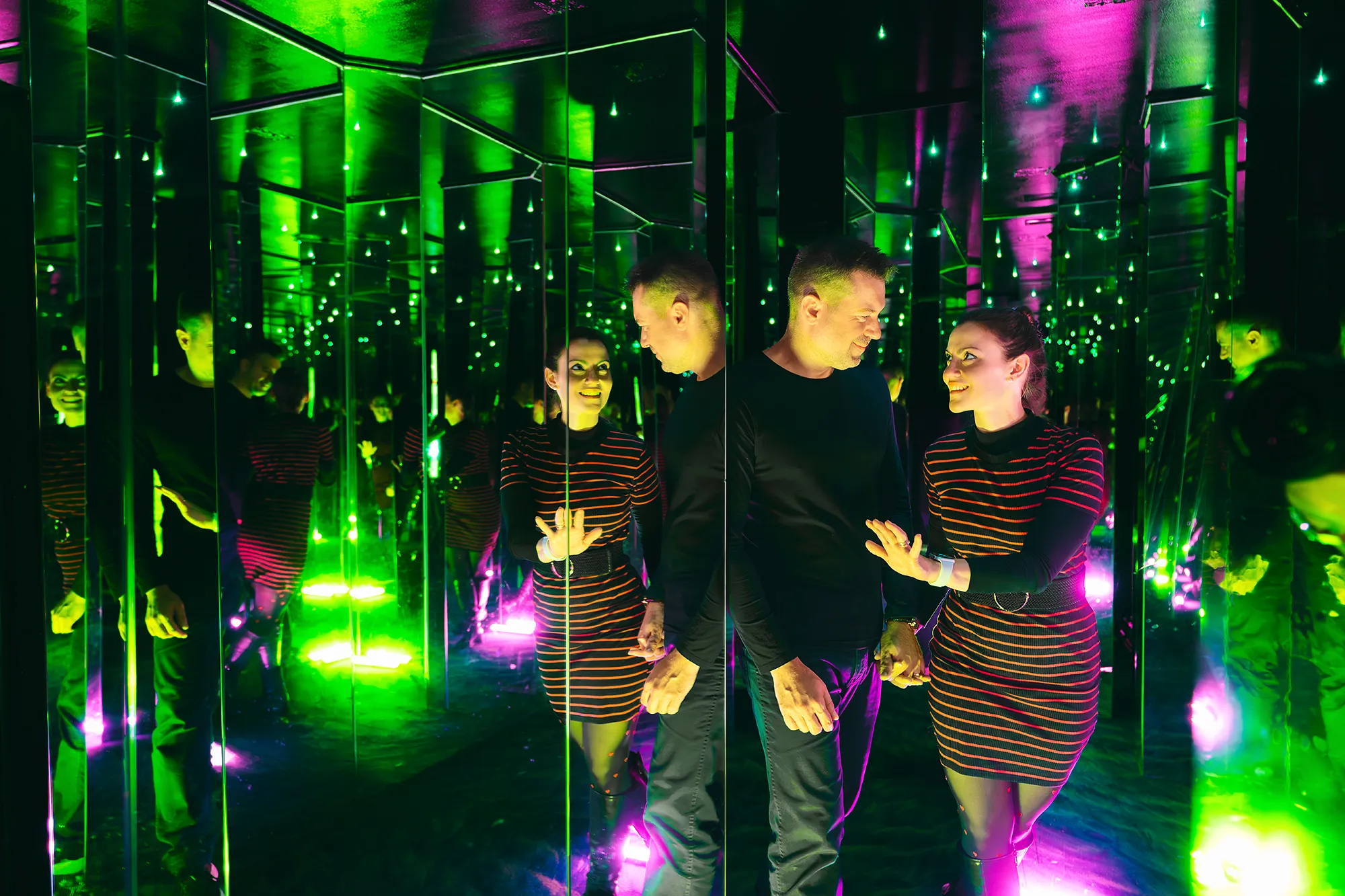 Two people standing in a mirrored room with green and purple lights creating multiple reflections