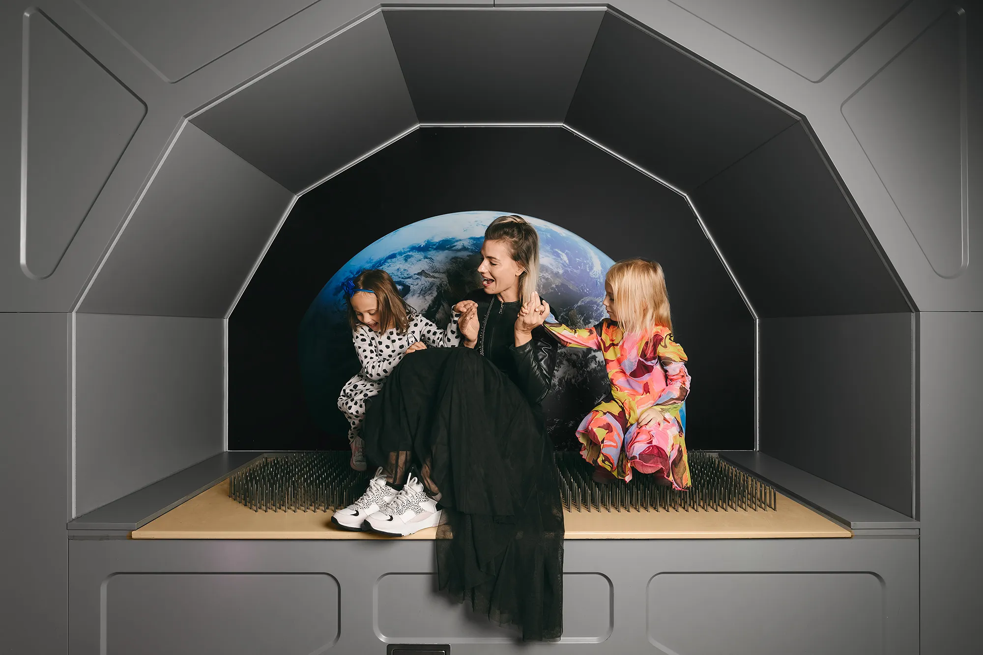 A woman and two children sitting inside a dark, futuristic pod with a large image of Earth in the background.