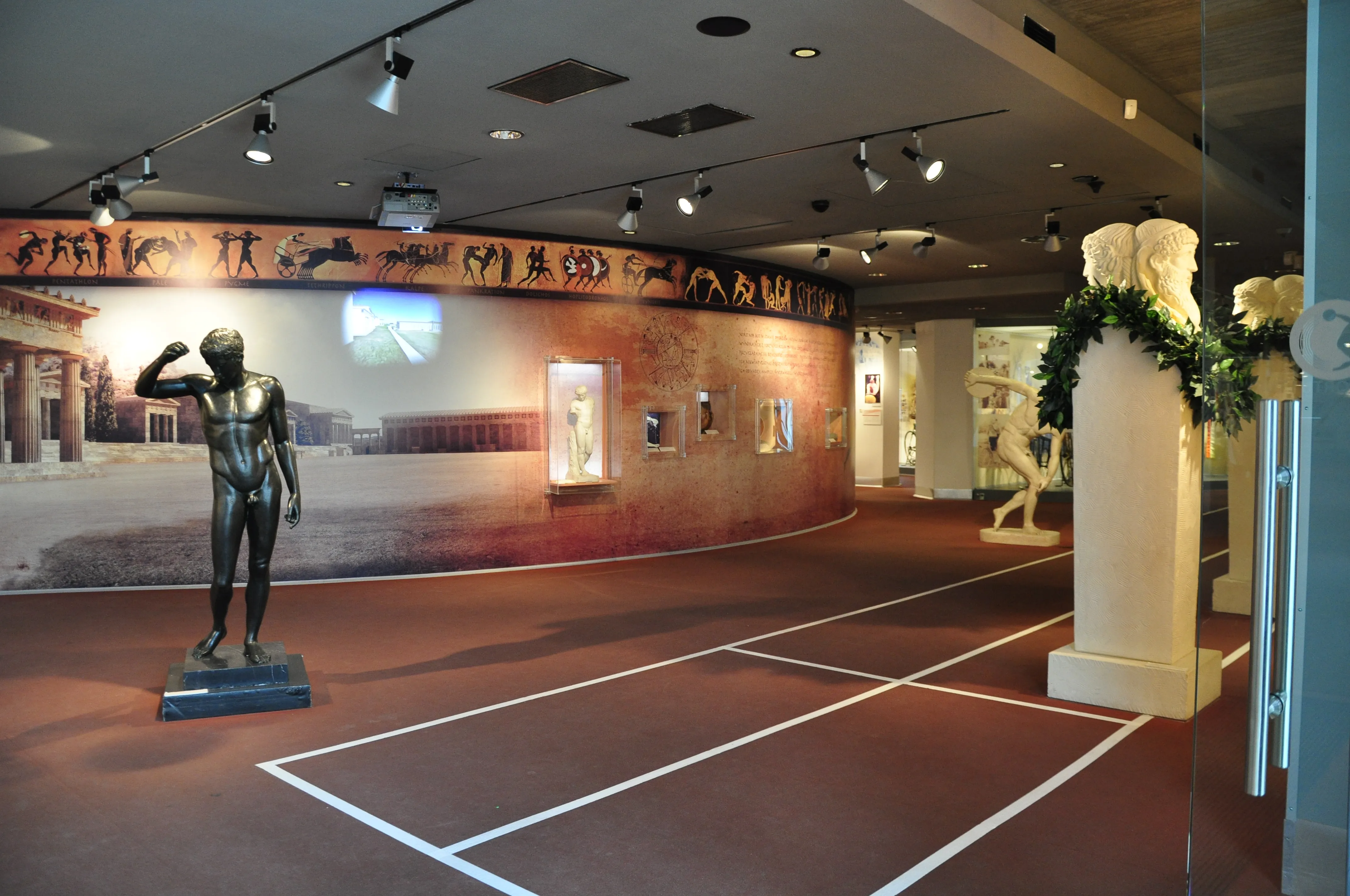 Interior of a museum with a bronze statue of an athlete, a running track floor, and a curved wall with murals and exhibits
