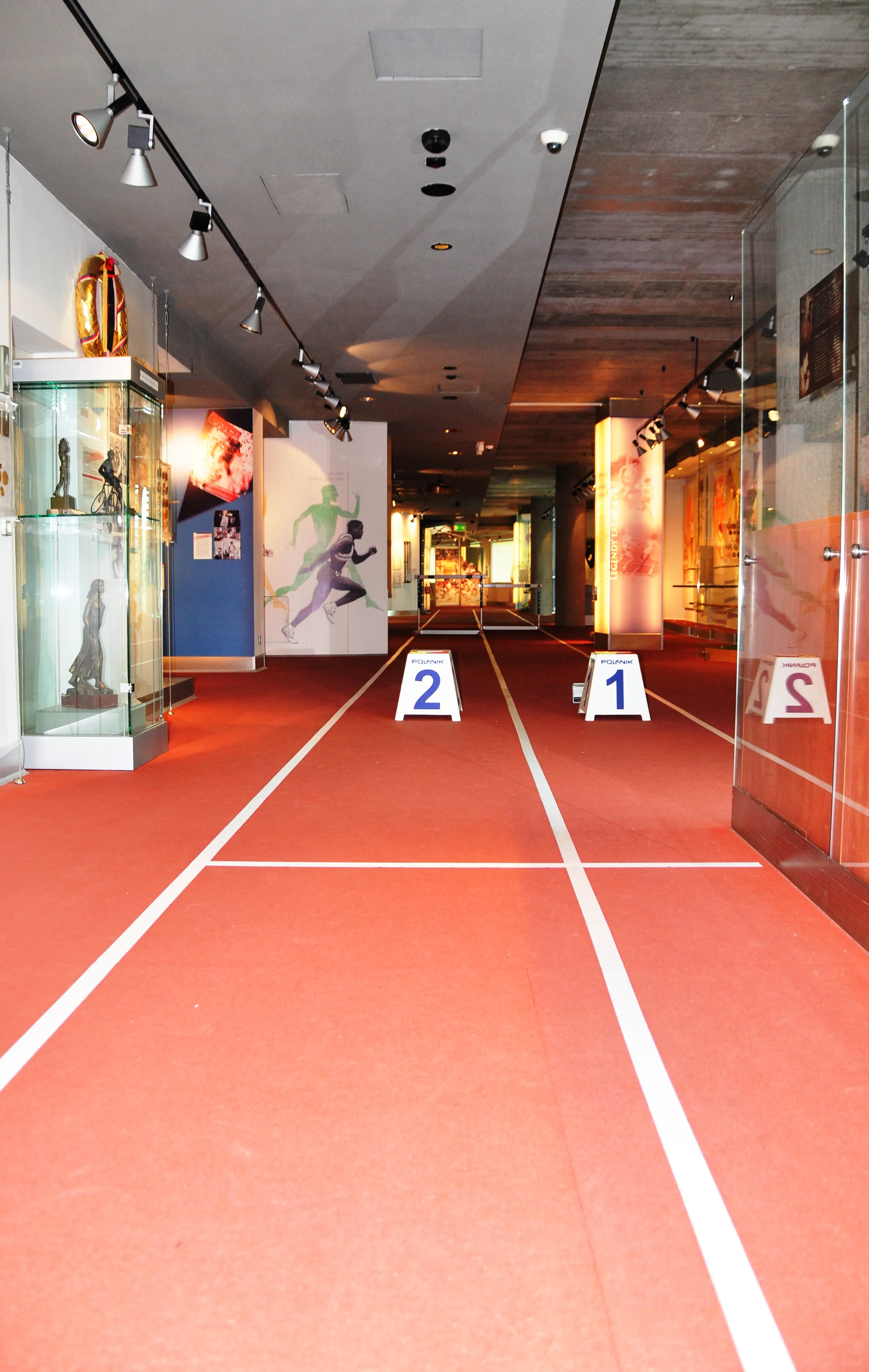 Interior hallway with red running track floor and white lane lines, display cases, and sports artwork