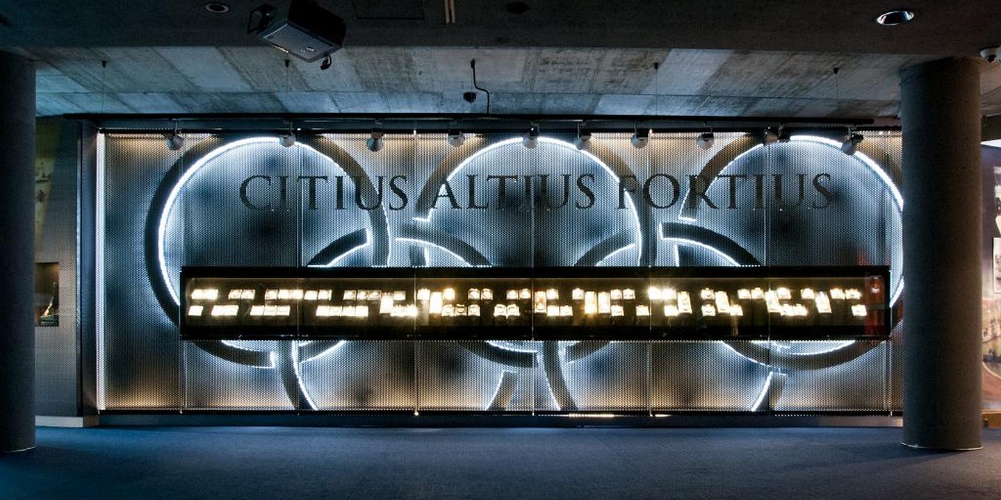 Illuminated display of the Olympic motto 'CITIUS ALTIUS FORTIUS' in a modern circular design with glowing rings and a lit display case below.