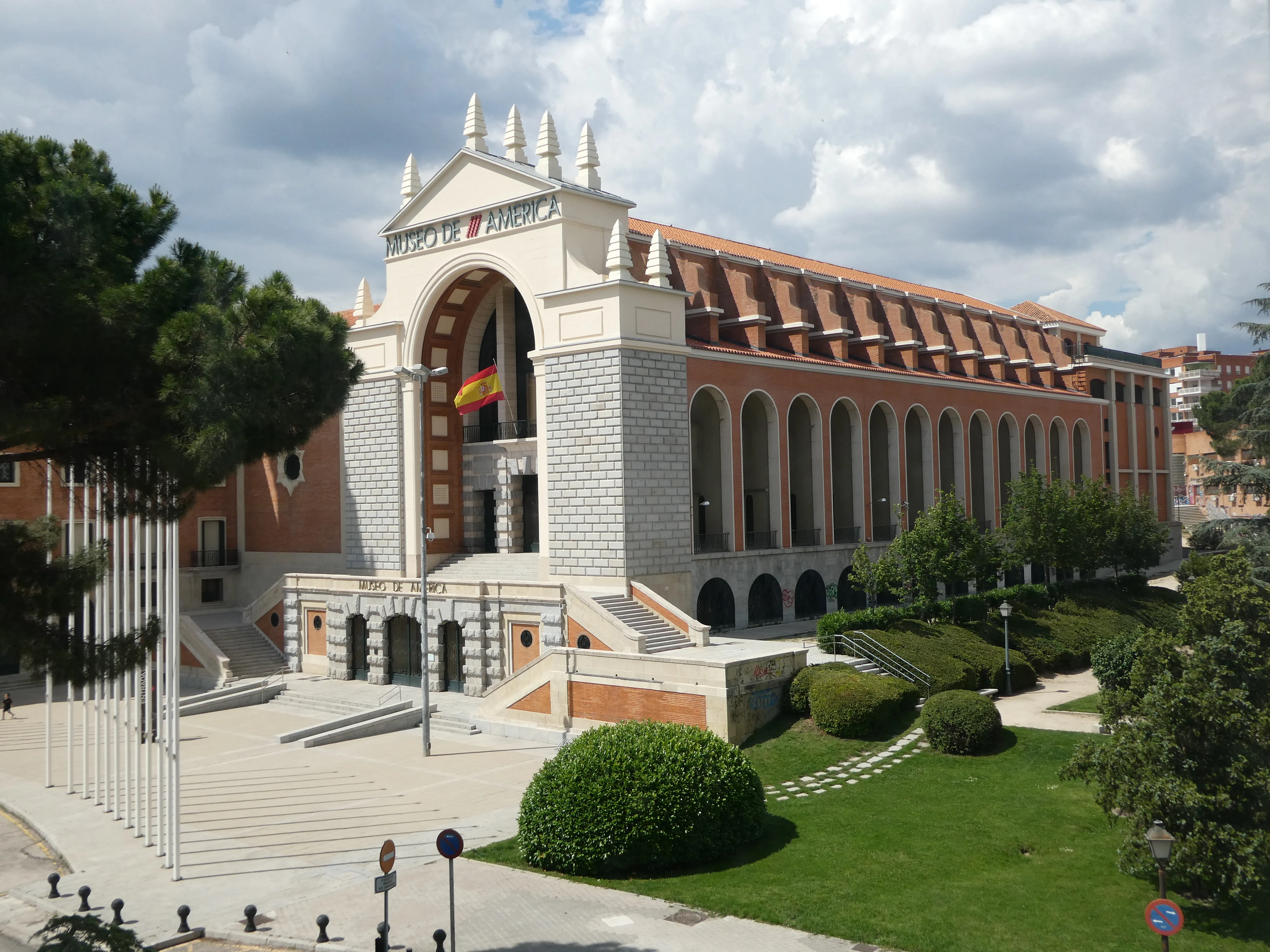 Zewnętrzna część Muzeum Ameryk w Madrycie z dużym łukowym wejściem, hiszpańską flagą i otaczającą zielenią