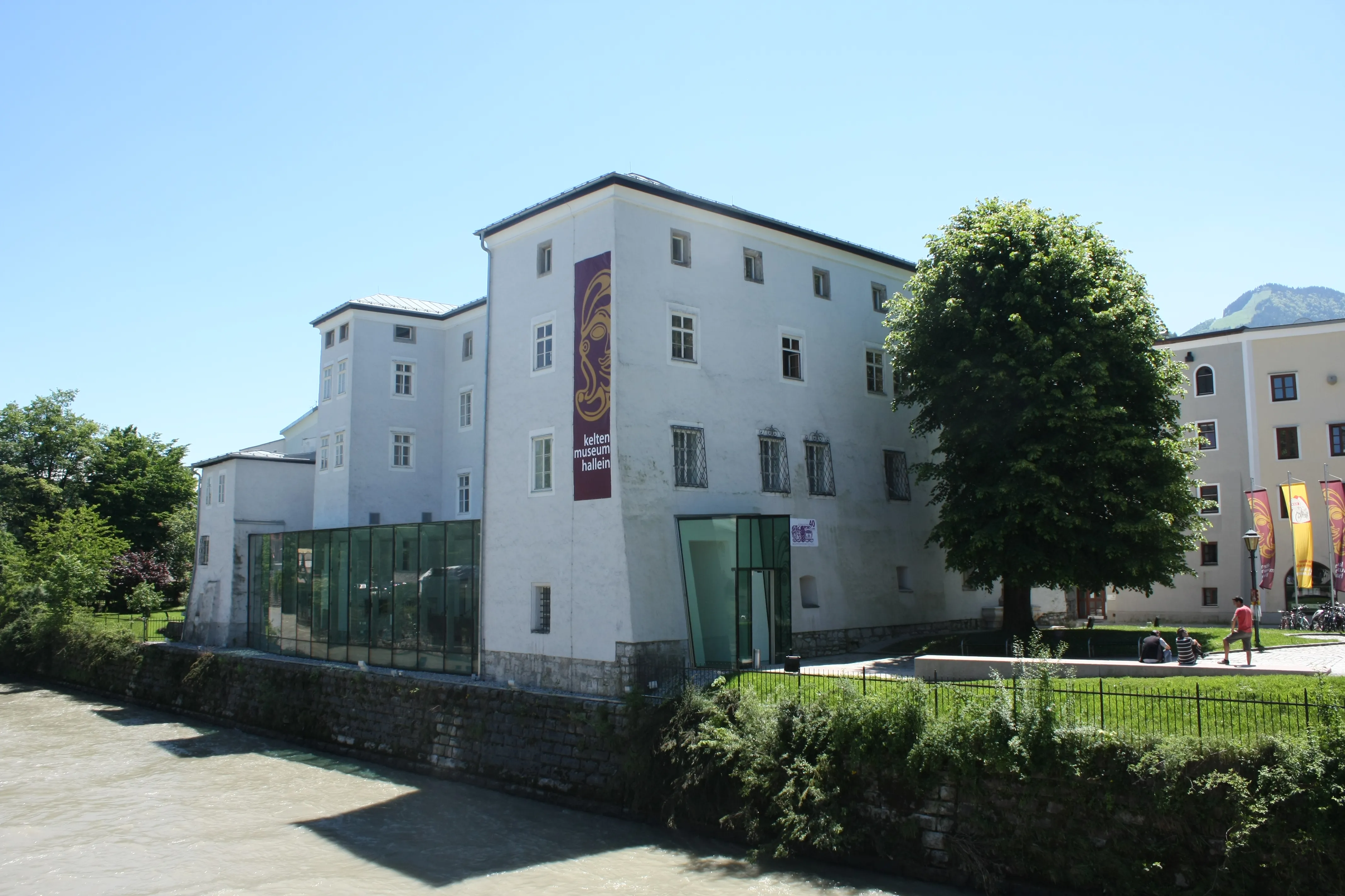 White multi-story museum building with a vertical banner reading 'Keltenmuseum' and a modern glass entrance extension, surrounded by green trees and hedges under a clear blue sky