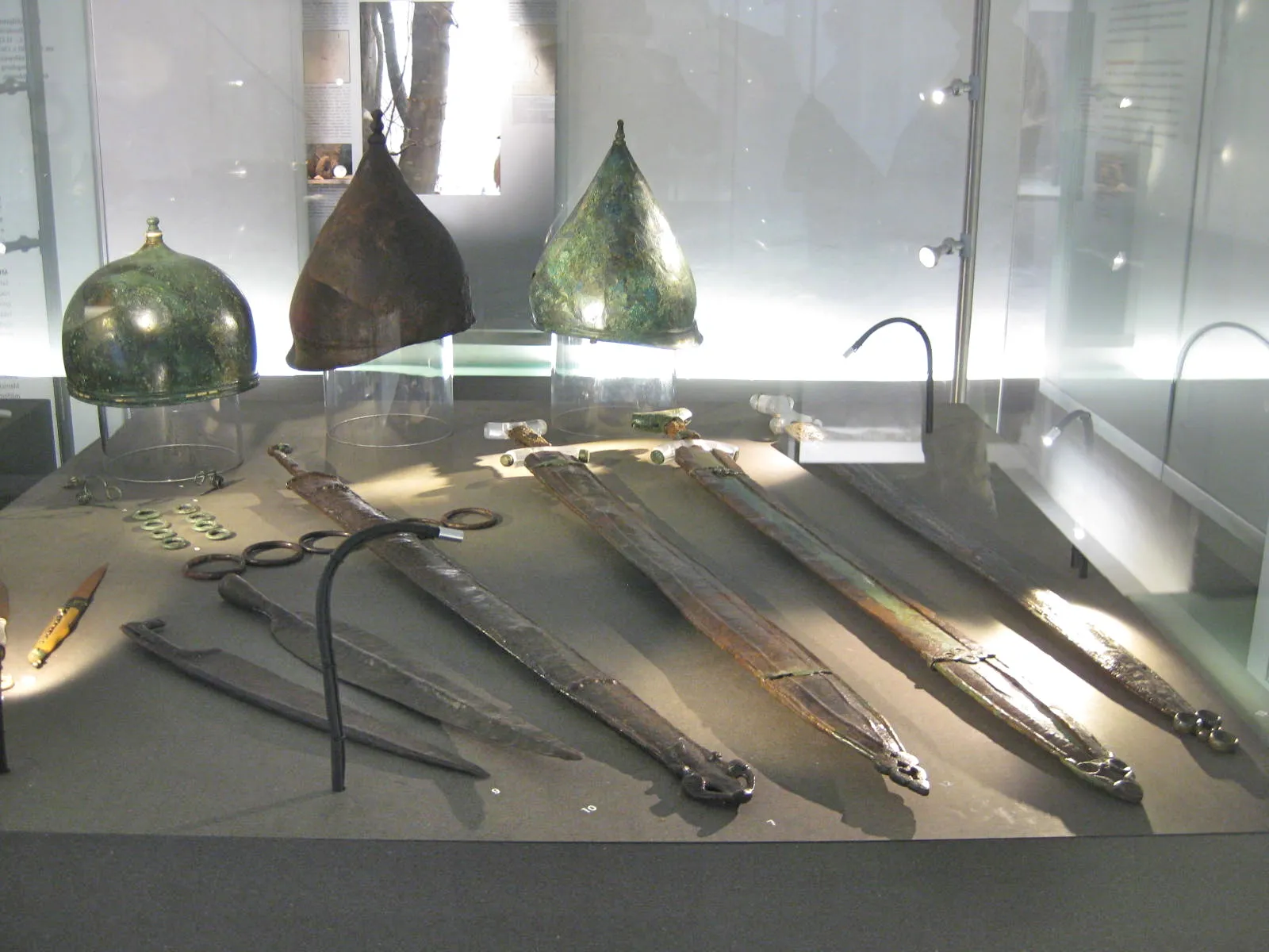 Display case containing three ancient Celtic helmets and several iron swords and daggers arranged on a gray surface.