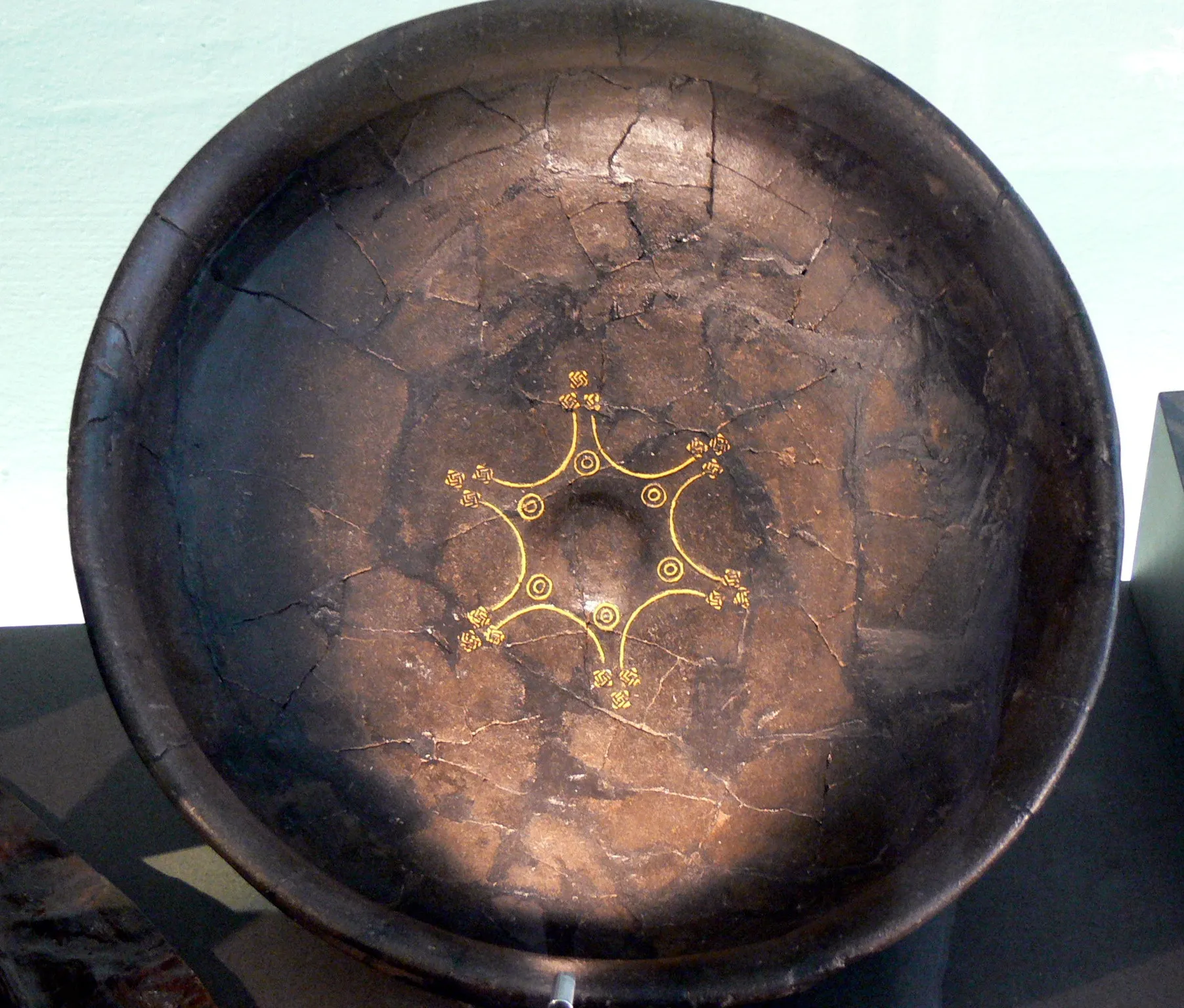 Ancient dark wooden bowl with a golden geometric pattern of circles and connecting lines at its center, photographed from above as a museum exhibit