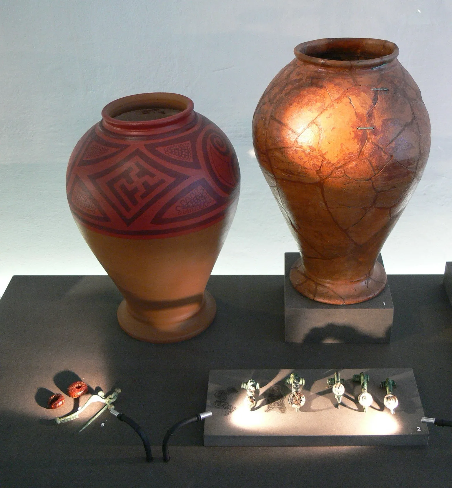 Dwa starożytne naczynia ceramiczne na podstawkach, oświetlone małymi reflektorami na wystawie muzealnej. Lewe naczynie zdobią czerwone wzory geometryczne, podczas gdy prawe ma popękaną, brązowawą powierzchnię.