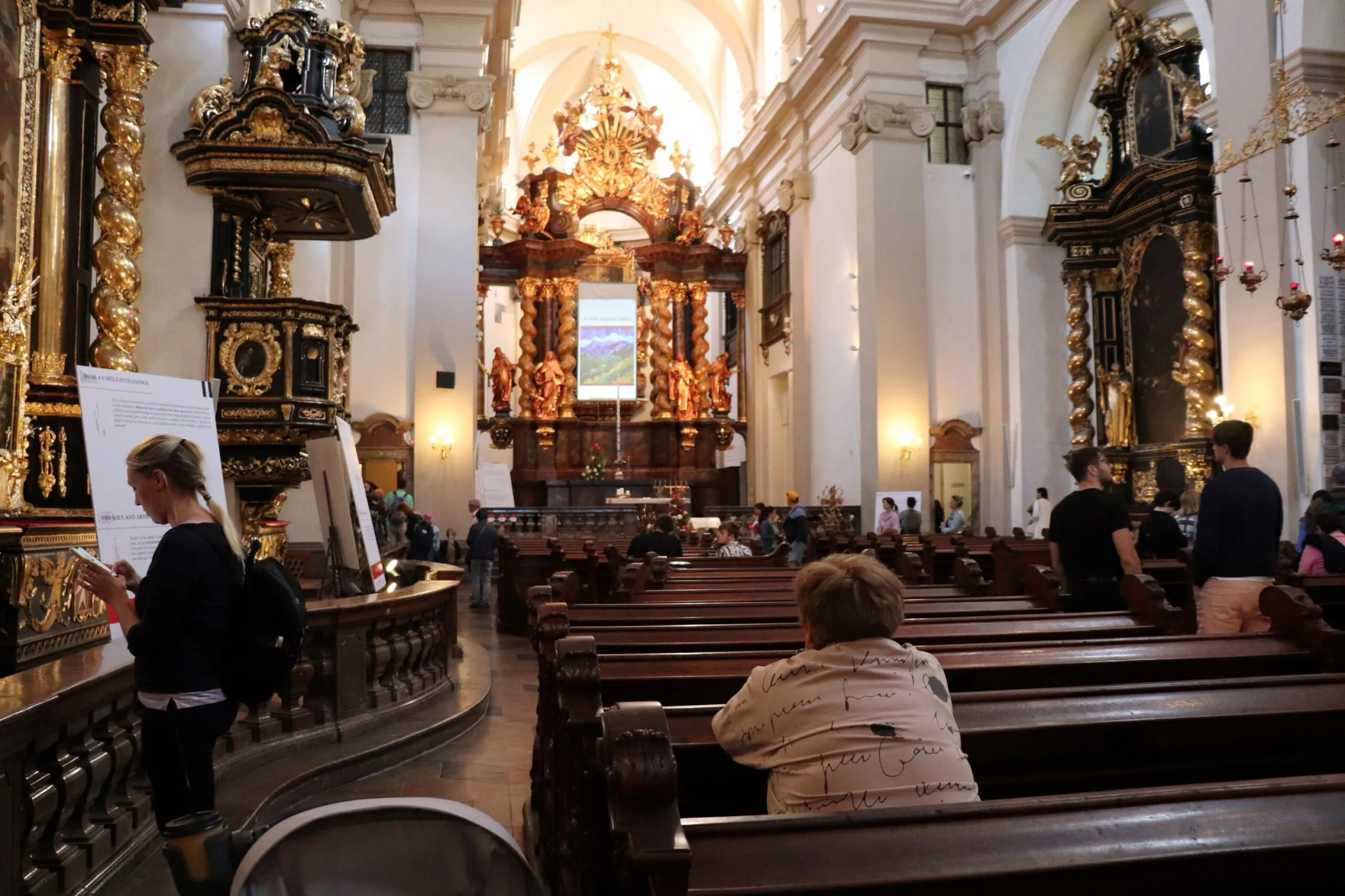 Interieur van een kerk met sierlijke gouden altaren, houten kerkbanken, en mensen die zitten en staan