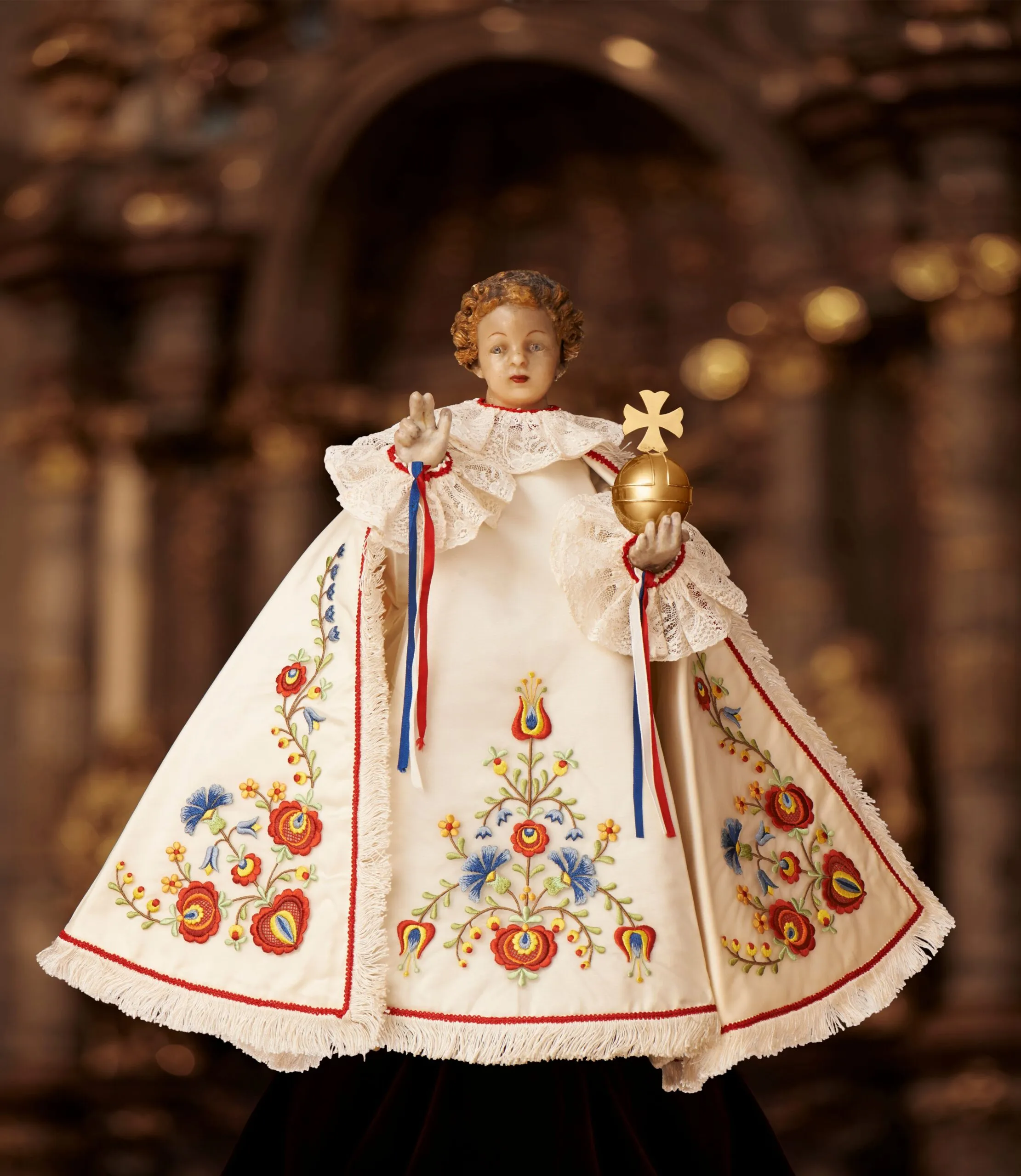 Woskowa figura Dzieciątka Jezus w ozdobnej białej szacie z kolorowymi haftami kwiatowymi, trzymająca złotą kulę i krzyż