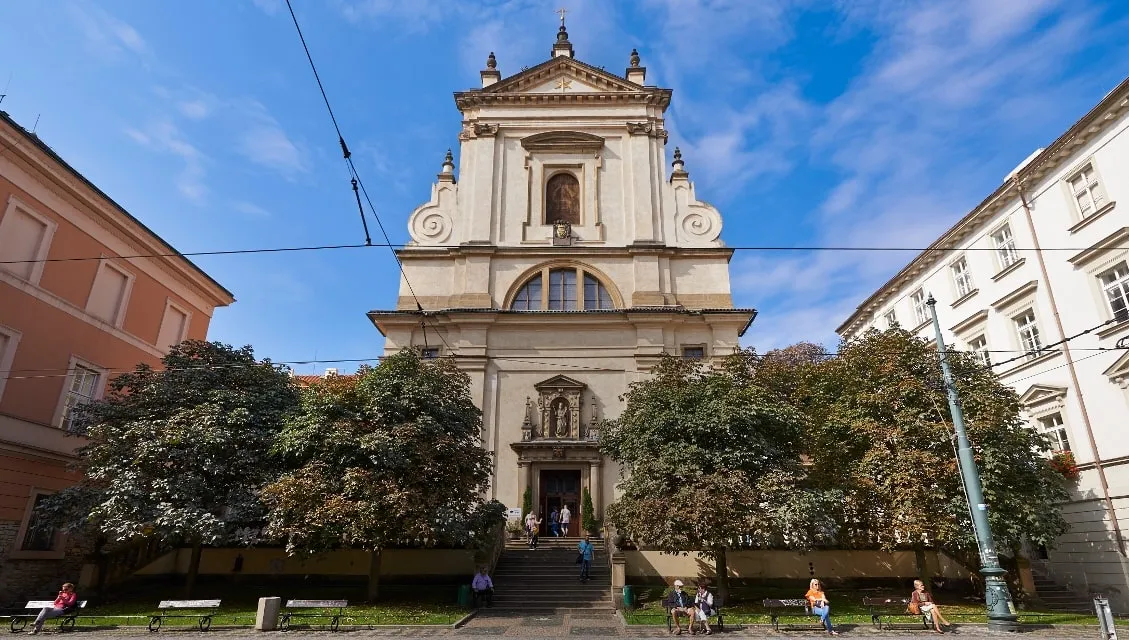 Fasada kościoła barokowego z centralnie umieszczoną rzeźbą, schodami prowadzącymi do wejścia i ludźmi siedzącymi na ławkach przed nim