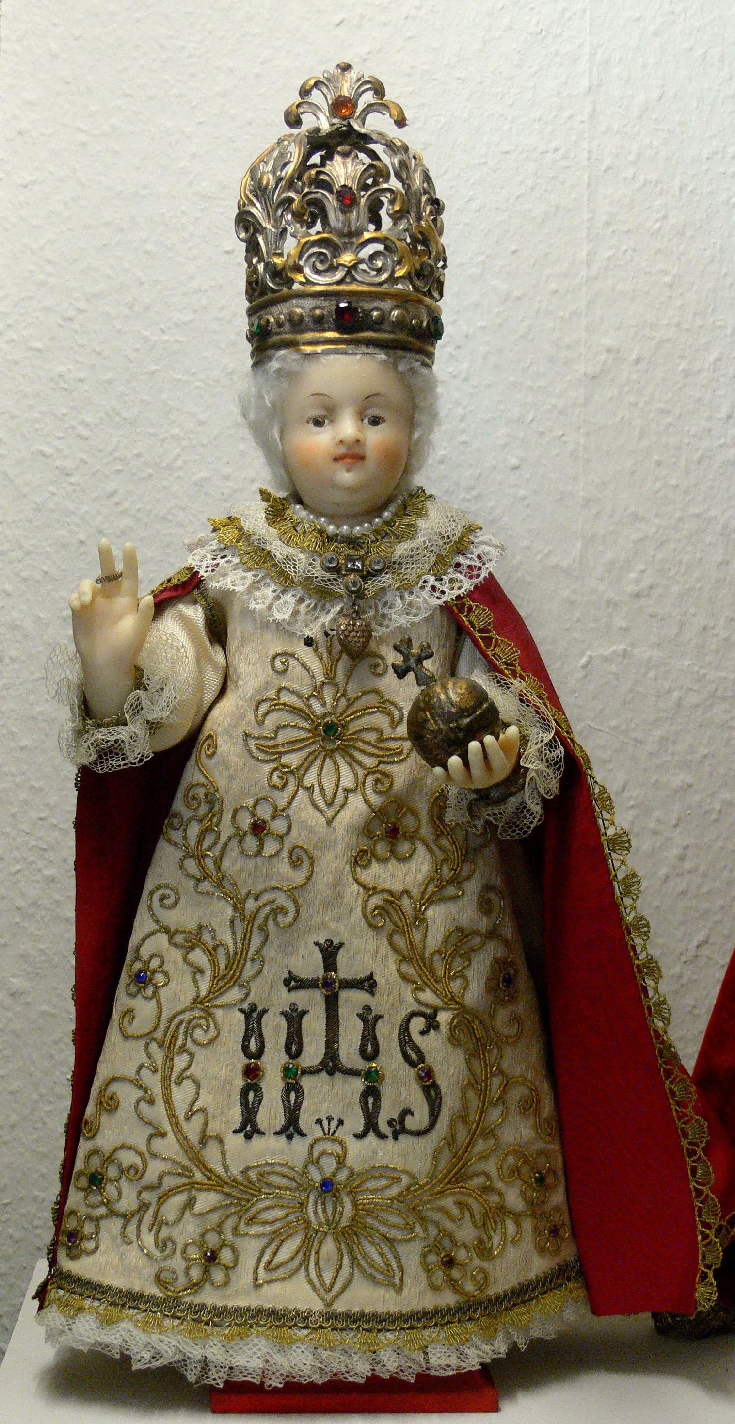 Woskowana figura Dzieciątka Jezus w złotej koronie, białej haftowanej szacie ze złotem i klejnotami oraz czerwonym płaszczu, trzymająca złote jabłko i wykonująca gest błogosławieństwa.