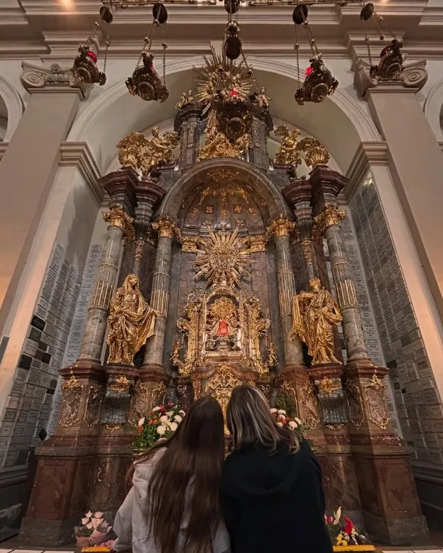 Twee mensen met lang haar die voor een groot, sierlijk, gouden altaar met een centraal beeld staan, omringd door gouden beelden en bloemen.
