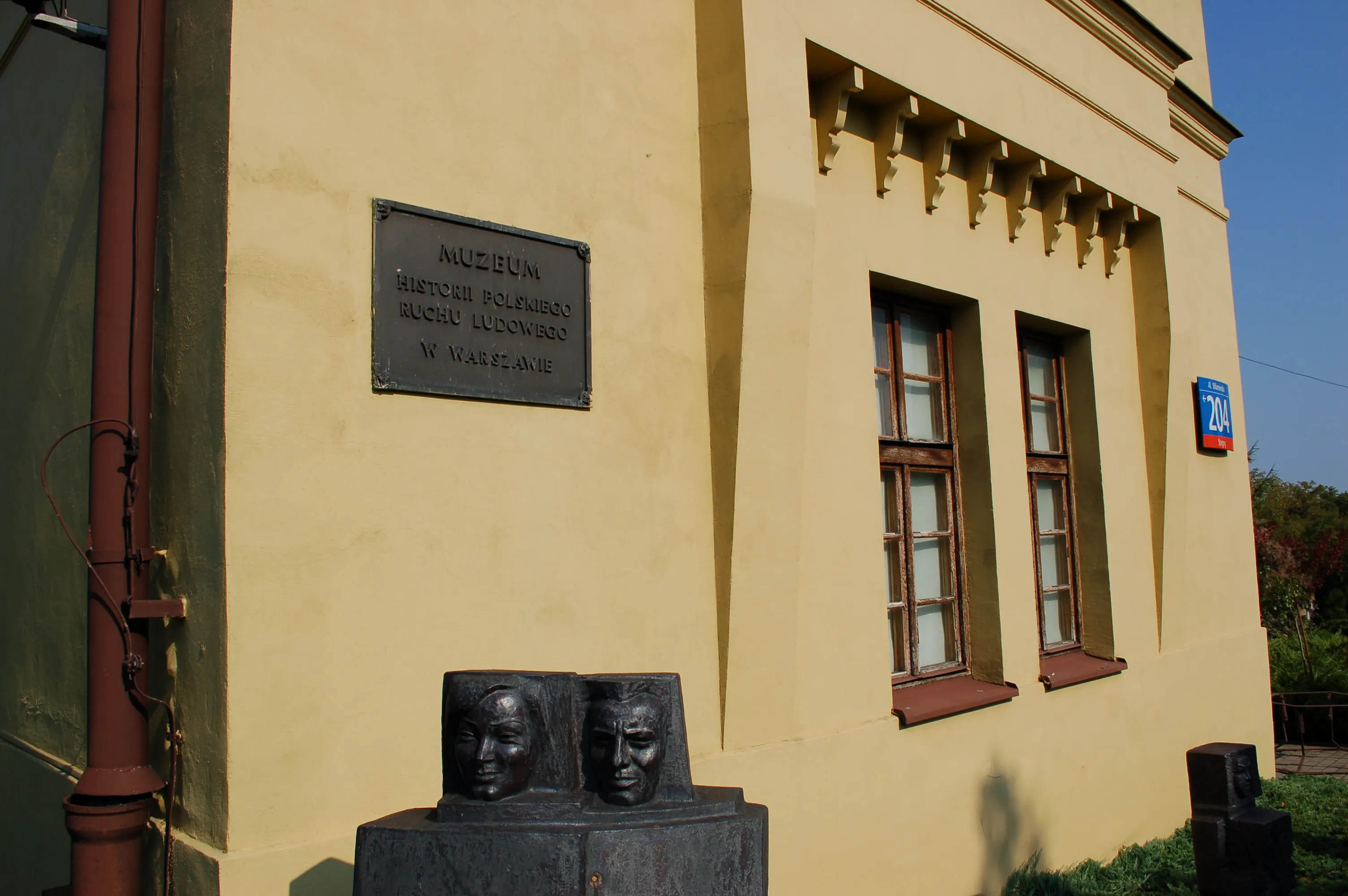 Żółty budynek z tablicą i rzeźbą dwóch twarzy z przodu