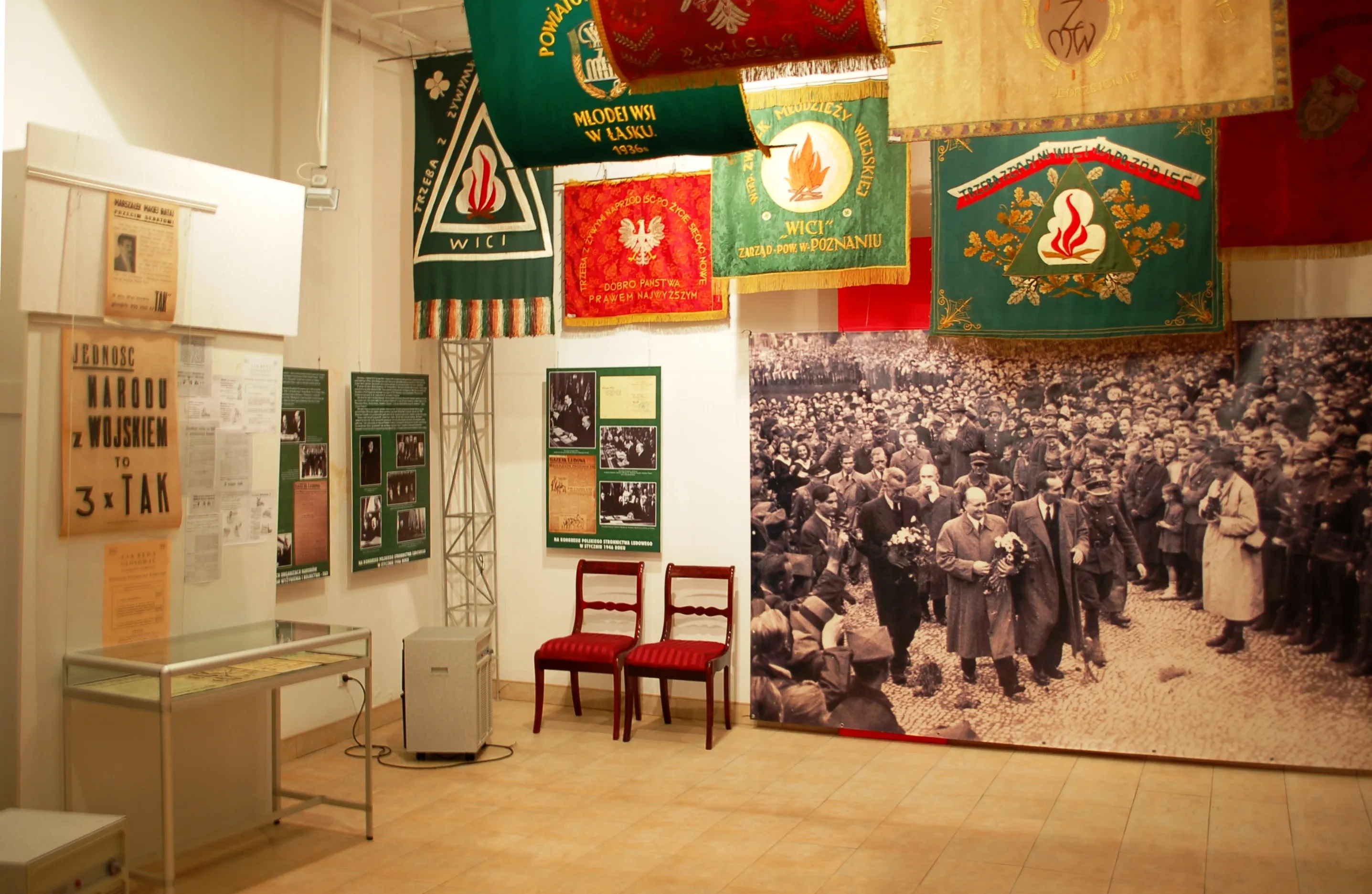 Wnętrze muzeum z historycznymi banerami, zdjęciem dużego tłumu i gablotami