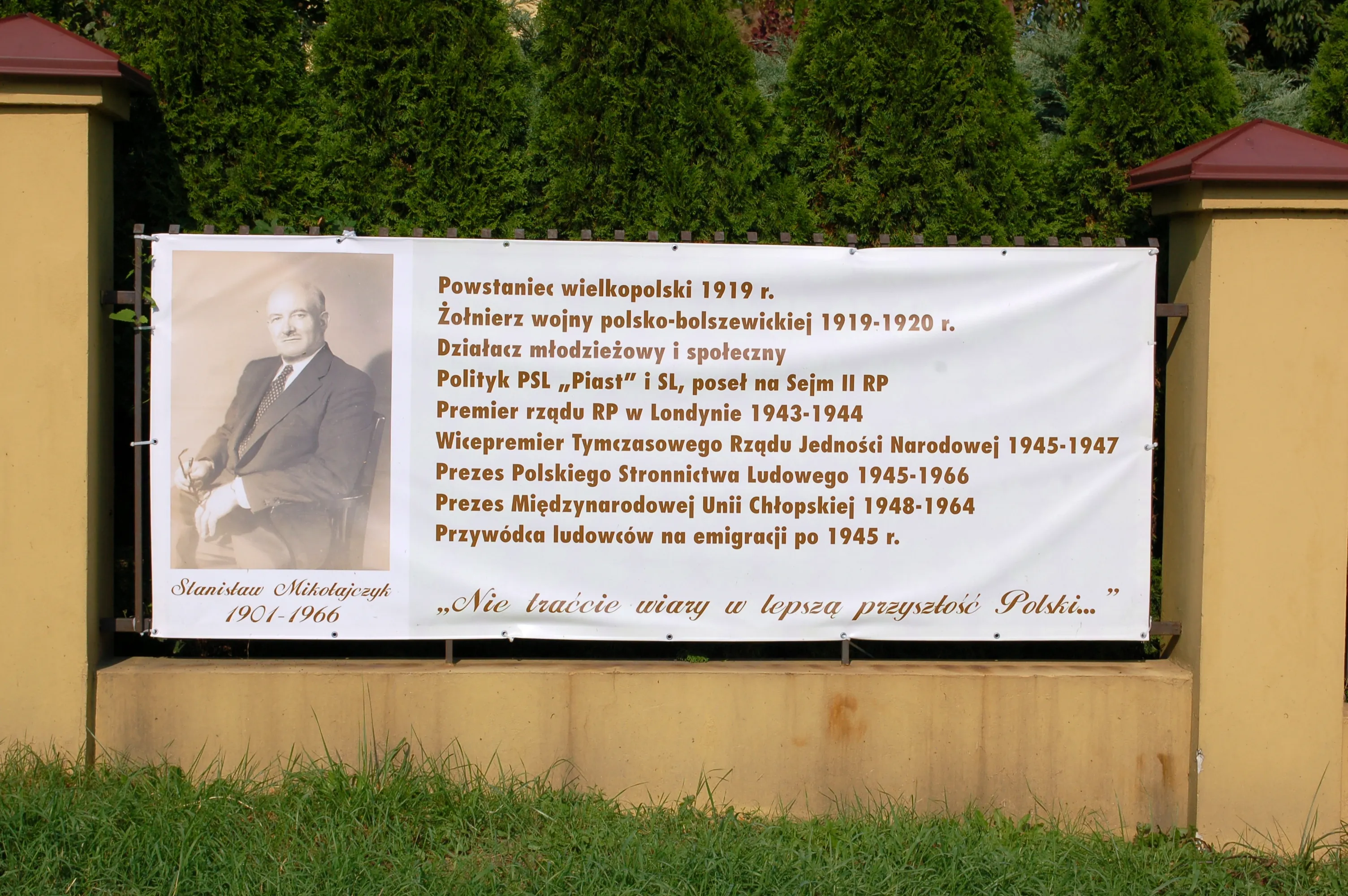 Een witte banier met een zwart-wit portret van een man en Poolse tekst die historische gebeurtenissen opsomt, gemonteerd aan een muur met groen gras en bomen op de achtergrond.