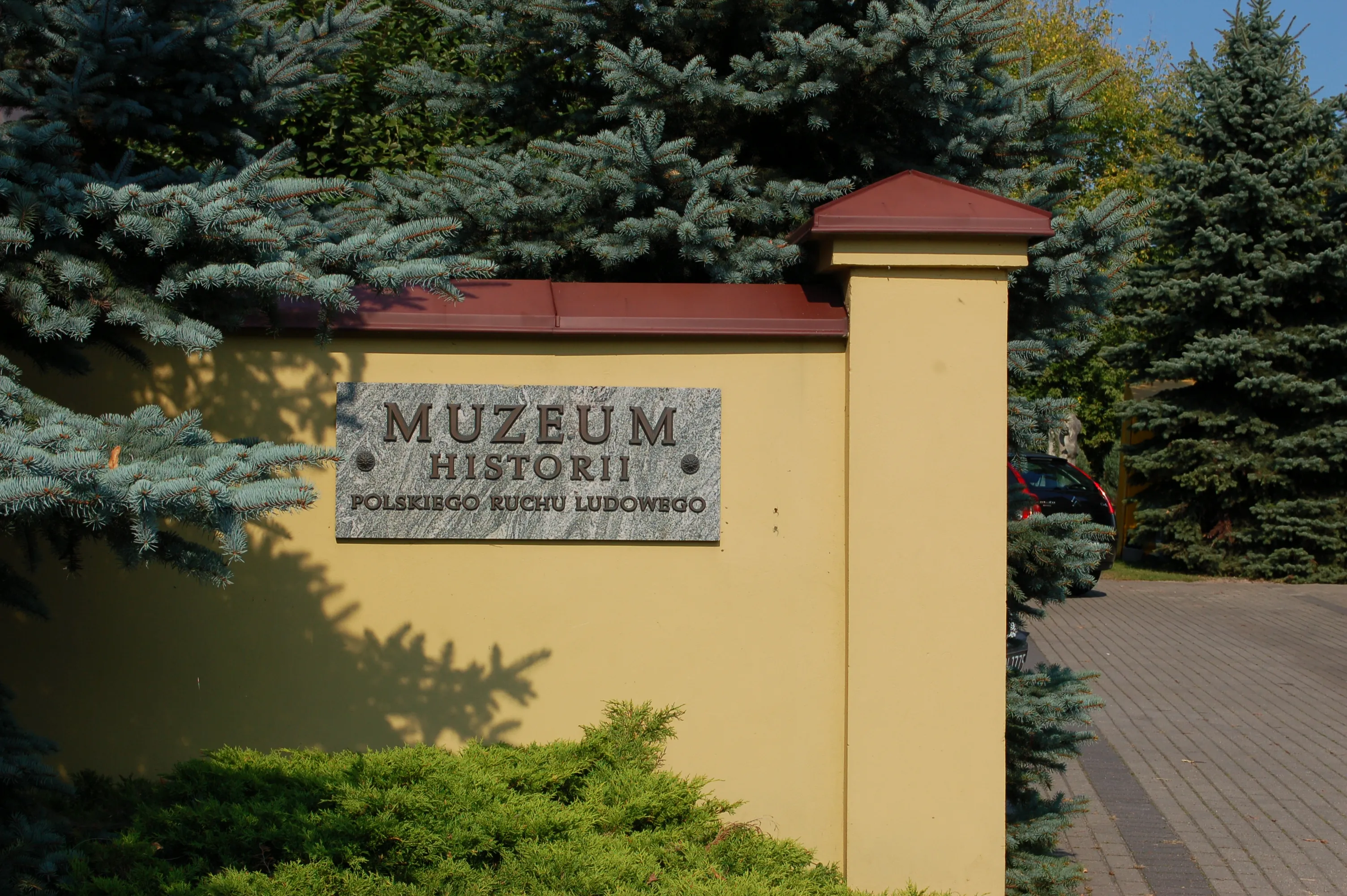 Kamienny napis 'MUZEUM HISTORII POLSKIEGO RUCHU LUDOWEGO' na żółtej ścianie z drzewami iglastymi
