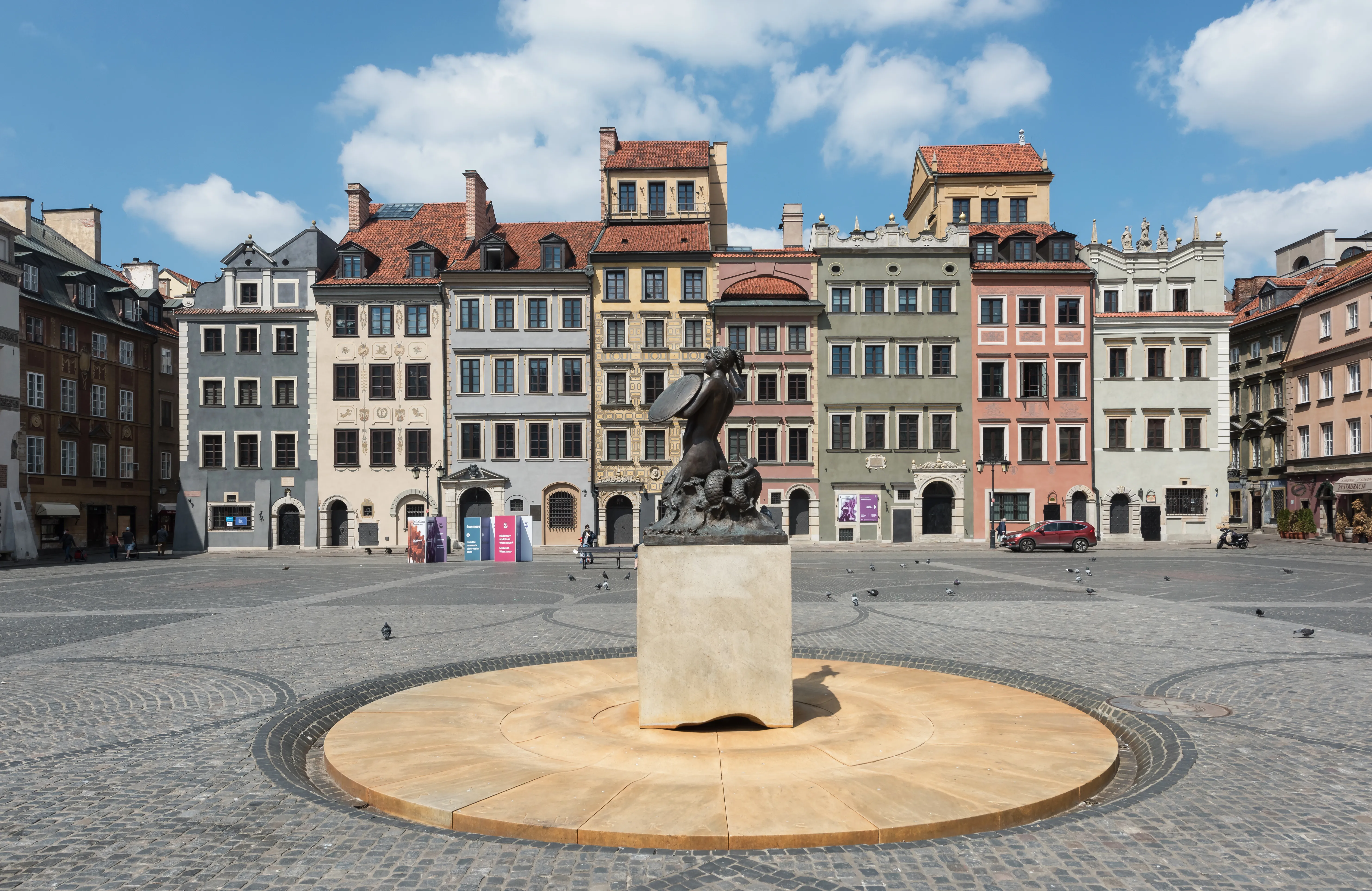 Het Syrenka-monument staat in het midden van een geplaveid plein, met historische gebouwen op de achtergrond onder een blauwe hemel.