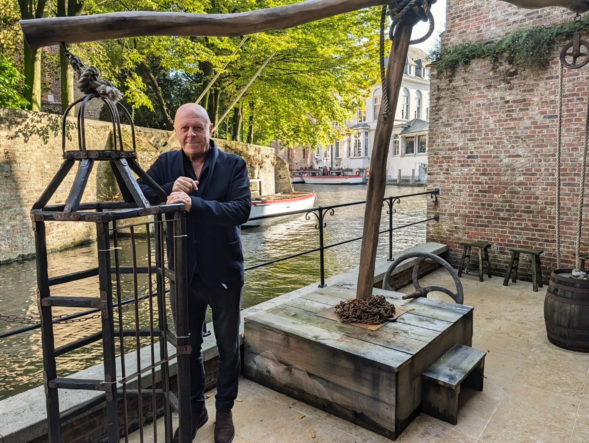 Un hombre está de pie junto a un gran mecanismo de grúa de madera con una estructura de jaula de hierro cerca de un canal, con edificios de ladrillo y árboles visibles al fondo.