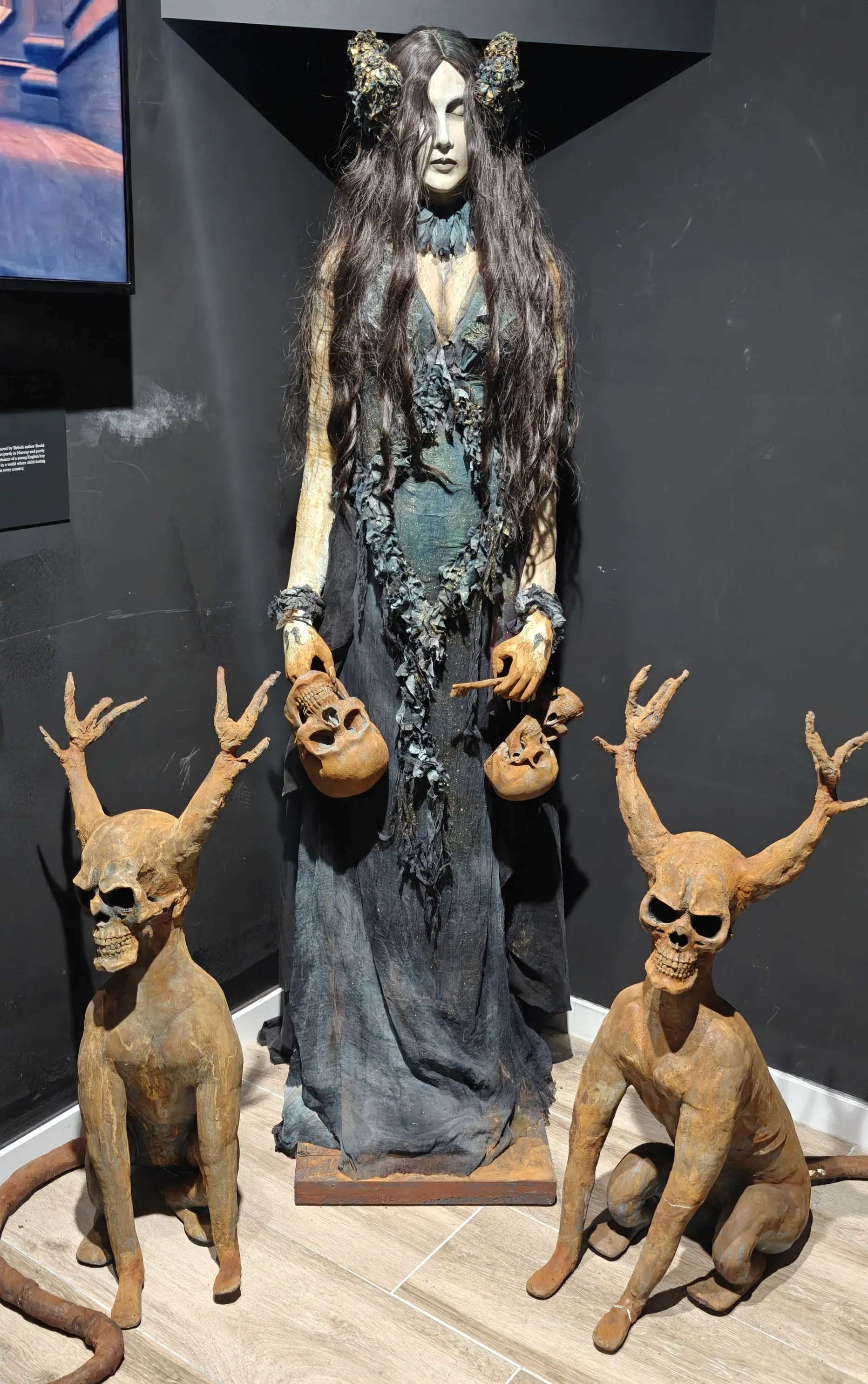 Exhibición de museo que presenta una figura femenina alta y pálida con un vestido oscuro y andrajoso que sostiene calaveras, flanqueada por dos criaturas esqueléticas con cuernos