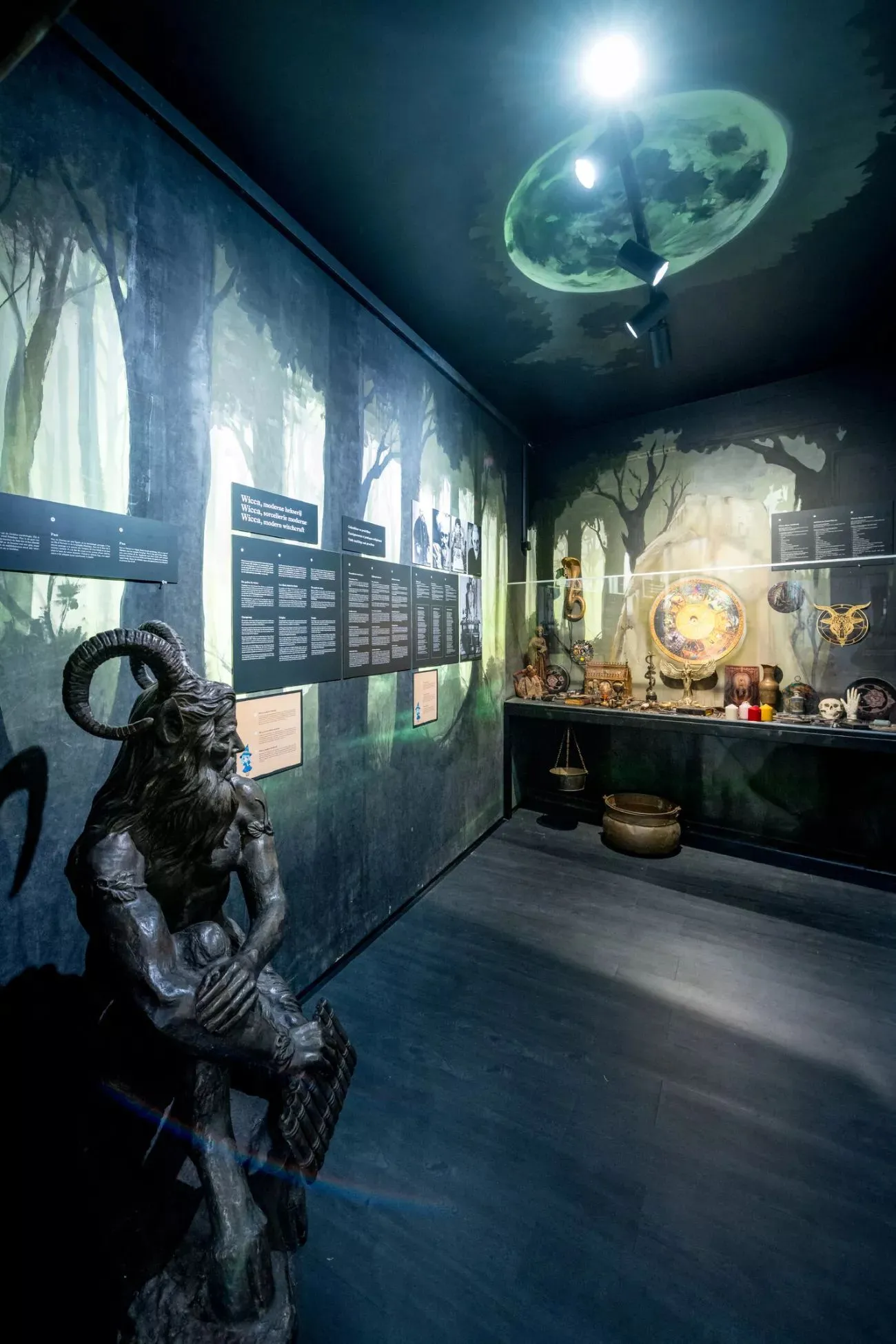 Sala de exposición del museo con iluminación tenue, un estatua con cuernos, murales de bosque, paneles informativos y una vitrina con objetos bajo una proyección de la luna en el techo.