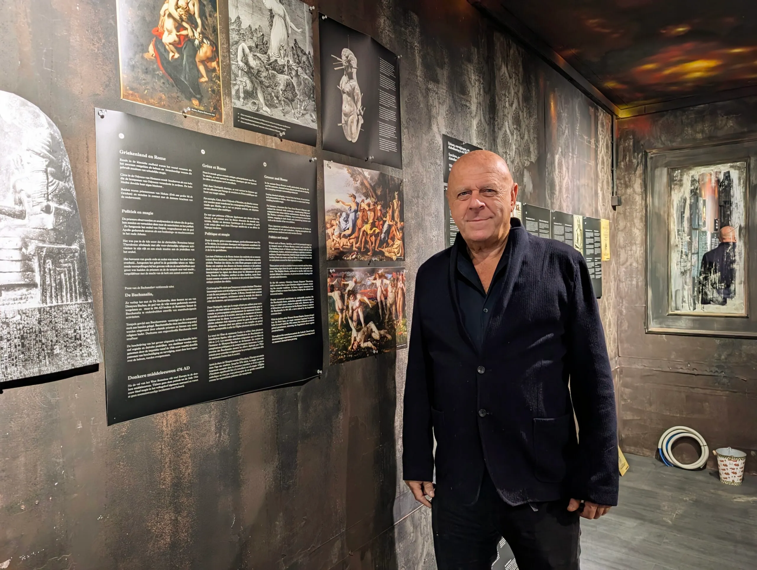 Un hombre con una americana oscura está de pie dentro de una galería de museo con paredes de madera oscura donde se exponen grabados artísticos históricos y paneles informativos sobre la brujería.