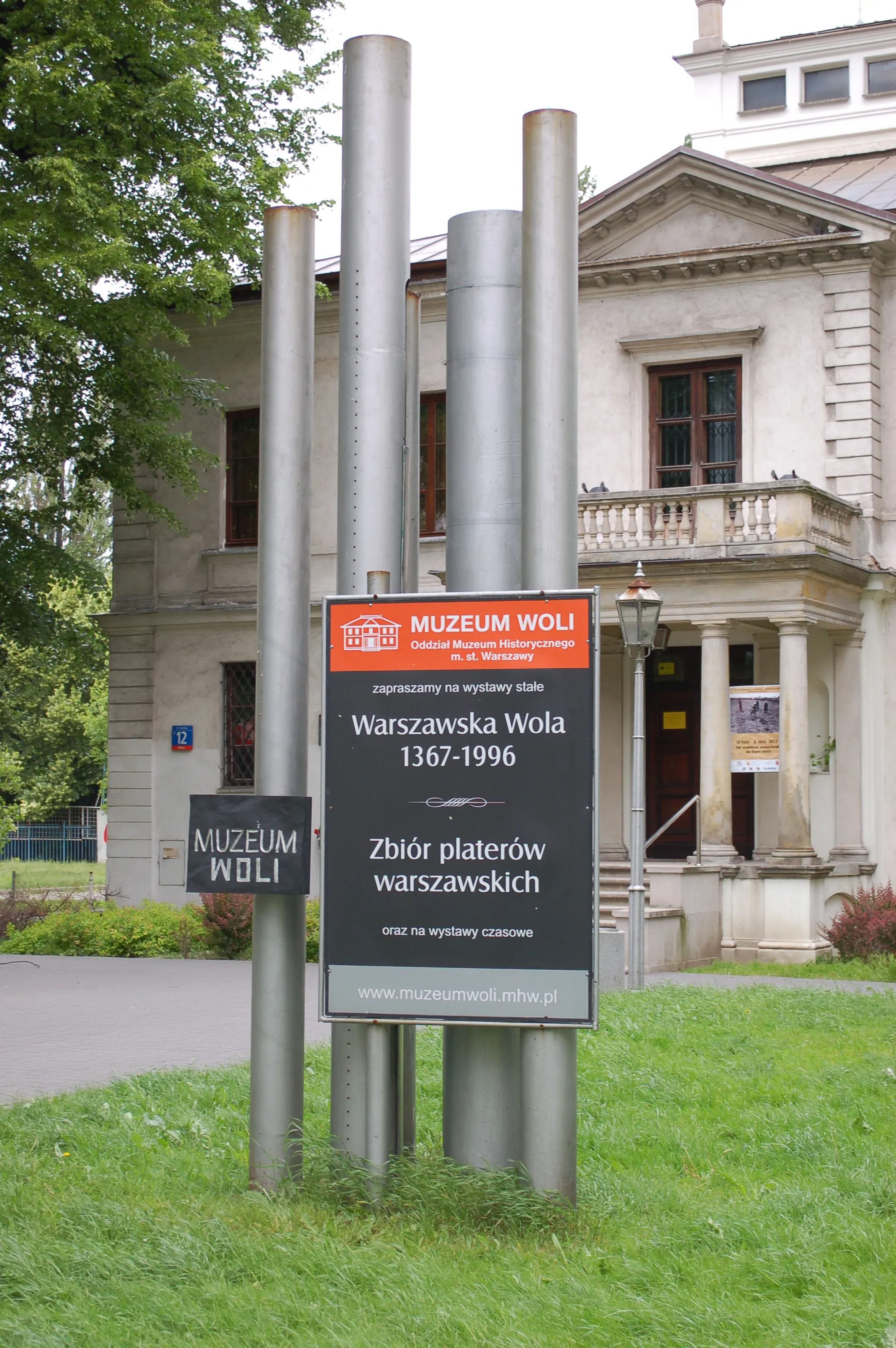 Znak Muzeum Woli stoi przed klasycystycznym budynkiem z kolumnami i balkonem, otoczonym zieloną trawą i drzewami.