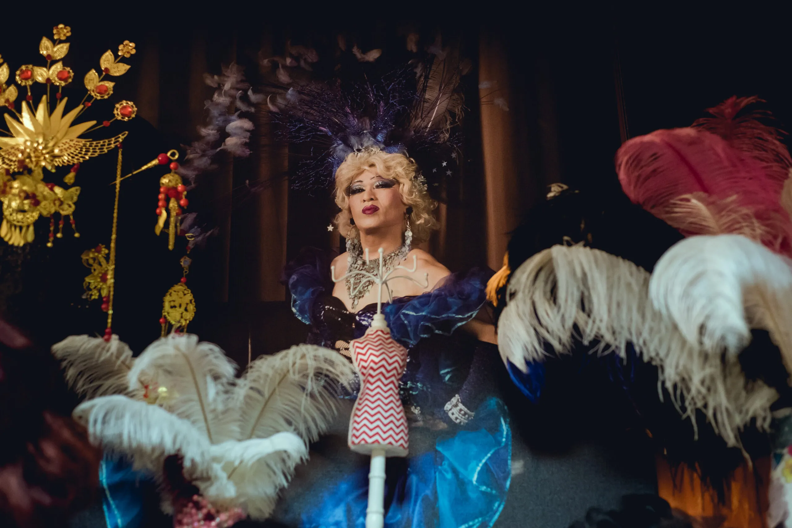 Una persona con un traje teatral y un tocado de plumas y un vestido azul está al lado de un maniquí con un top estampado, rodeada de ornamentadas decoraciones doradas y plumas.