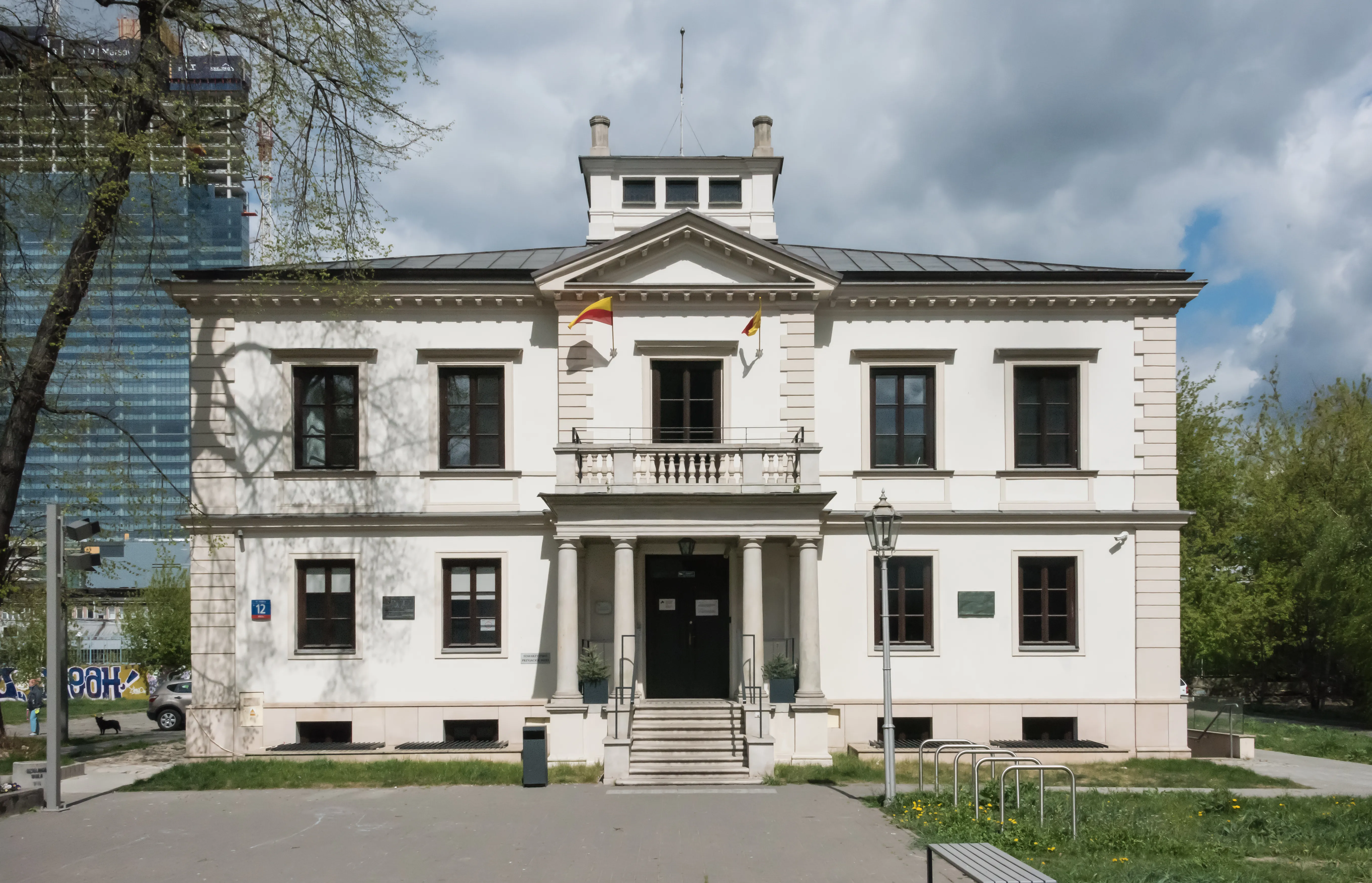 Biały budynek w stylu neoklasycystycznym z centralnym wejściem, balkonem i wieloma oknami pod zachmurzonym niebem