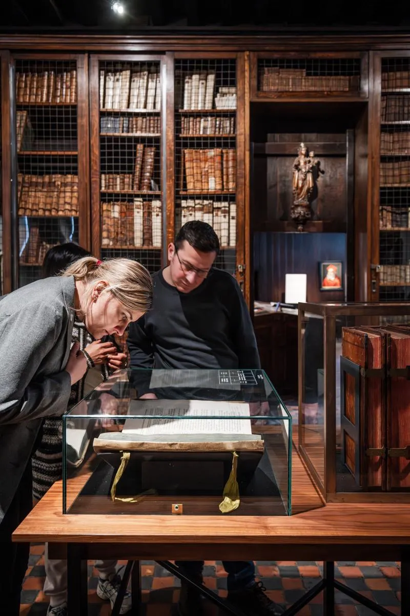 Dos visitantes se inclinan sobre una vitrina de cristal para examinar un libro abierto, con altas estanterías de madera llenas de libros antiguos al fondo.