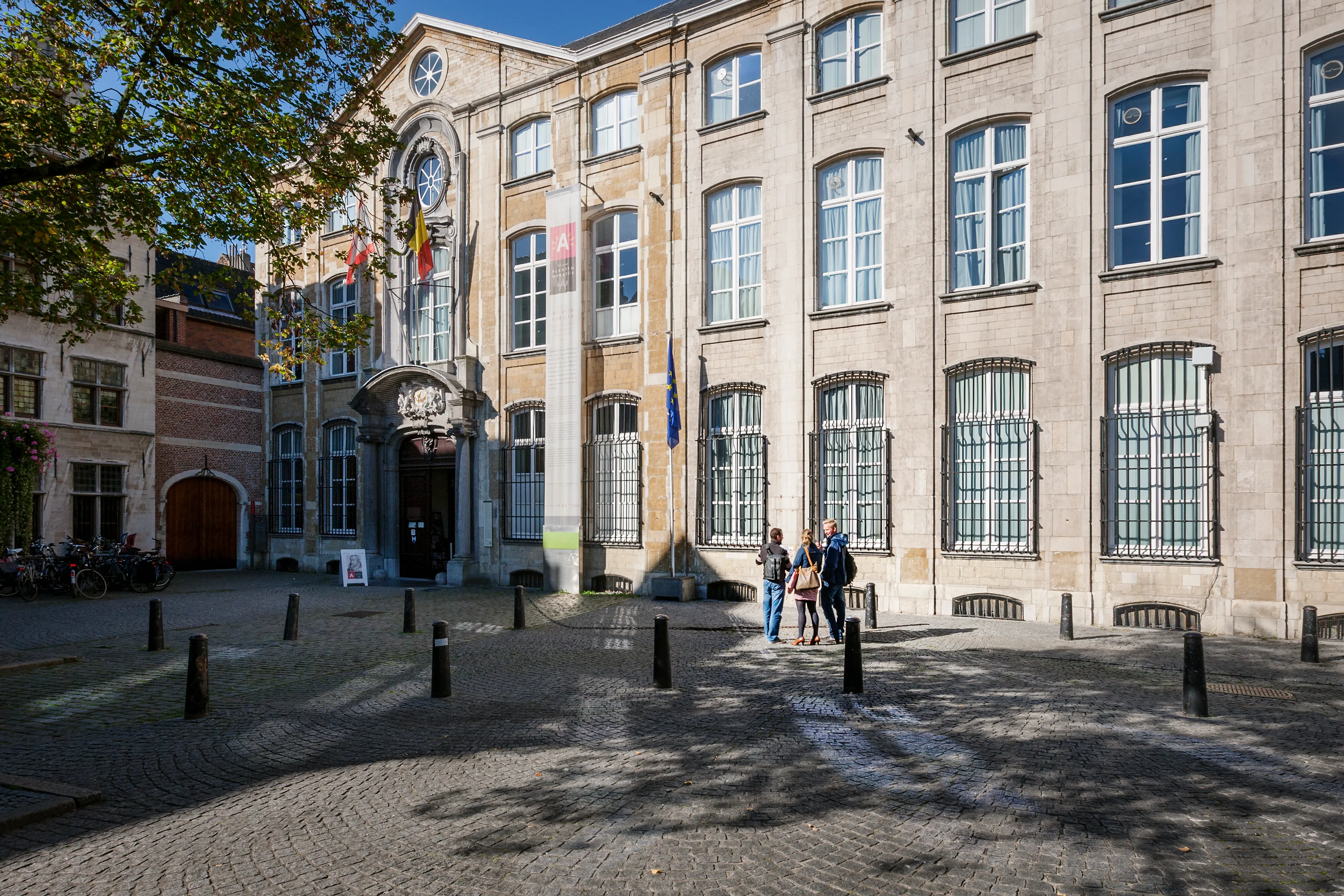 Stone facade of Museum Plantin-Moretus with arched entrance, flags, and cobblestone courtyard with visitors