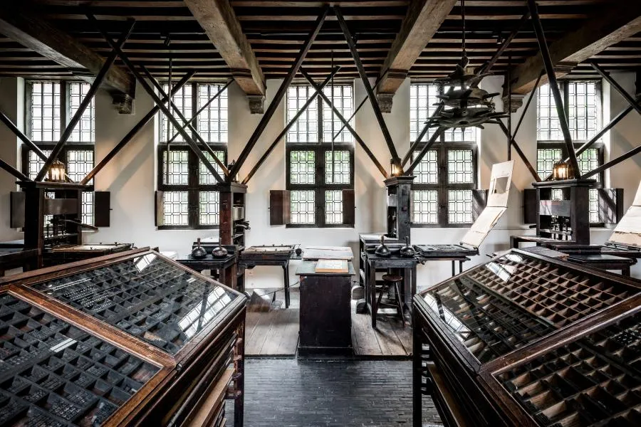 Interior del Museo Plantin-Moretus que muestra prensas de imprimir históricas y cajas de tipos en una sala con grandes ventanales y vigas de madera expuestas en el techo