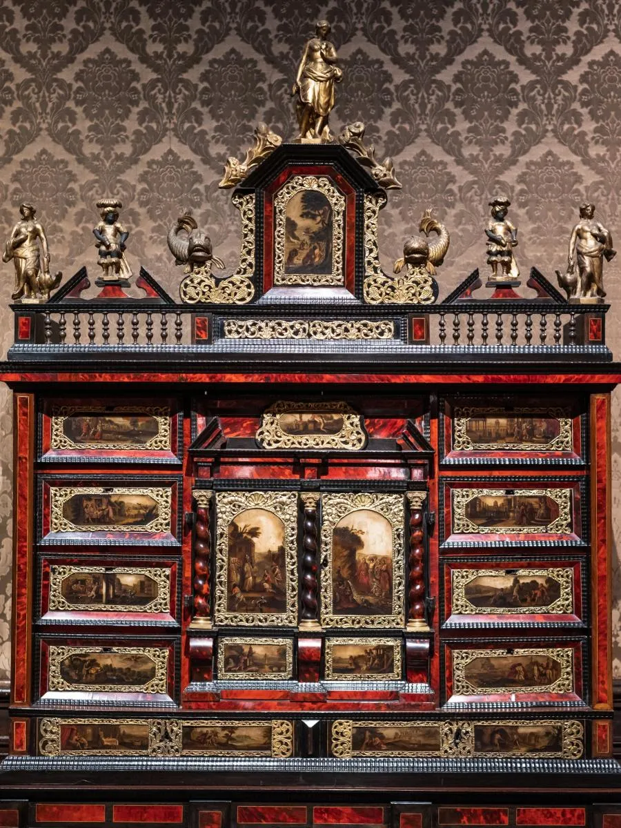 Un gabinete ornamentado de varios cajones con laca roja y negra, bordes dorados, pequeños paneles pintados y figuritas de bronce en la parte superior, colocado sobre un papel tapiz estampado.