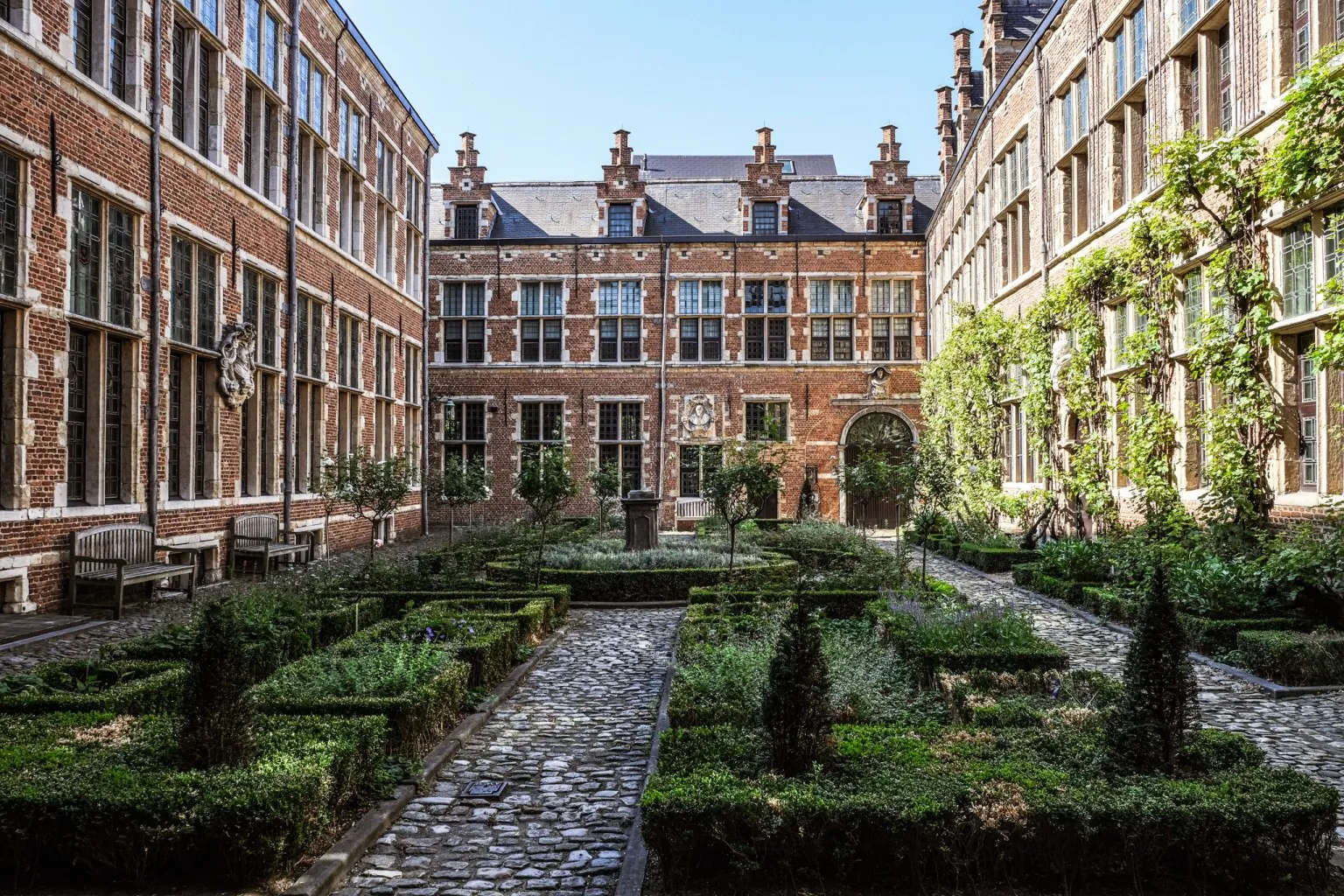 Un jardín formal con setos cuidadosamente recortados, caminos de adoquines e históricos edificios de ladrillo con grandes ventanales que rodean el espacio verde.